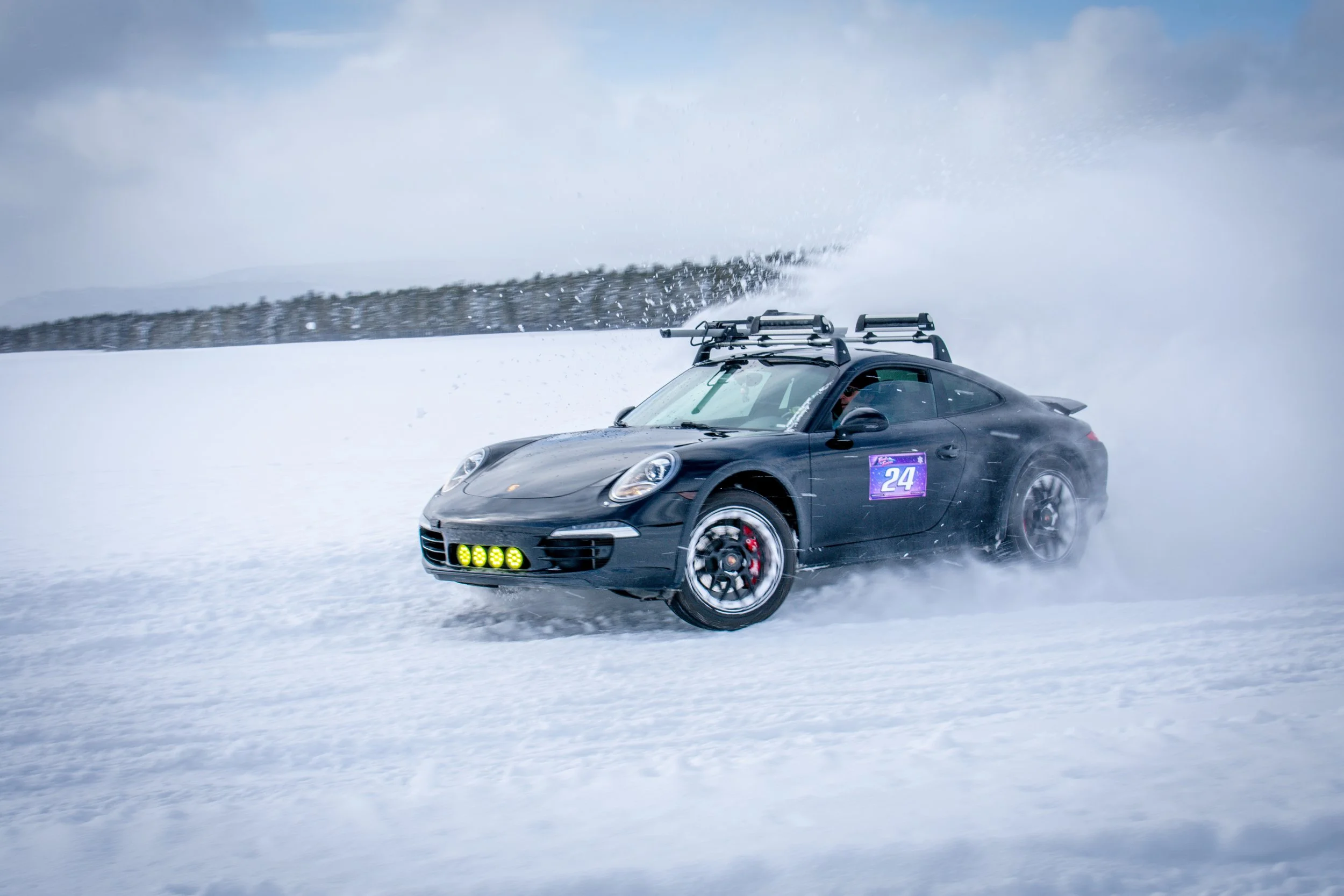 Black sports car with a number 24 racing label driving on snow, creating a spray of snow behind it, with a background of snow-covered landscape and cloudy sky.