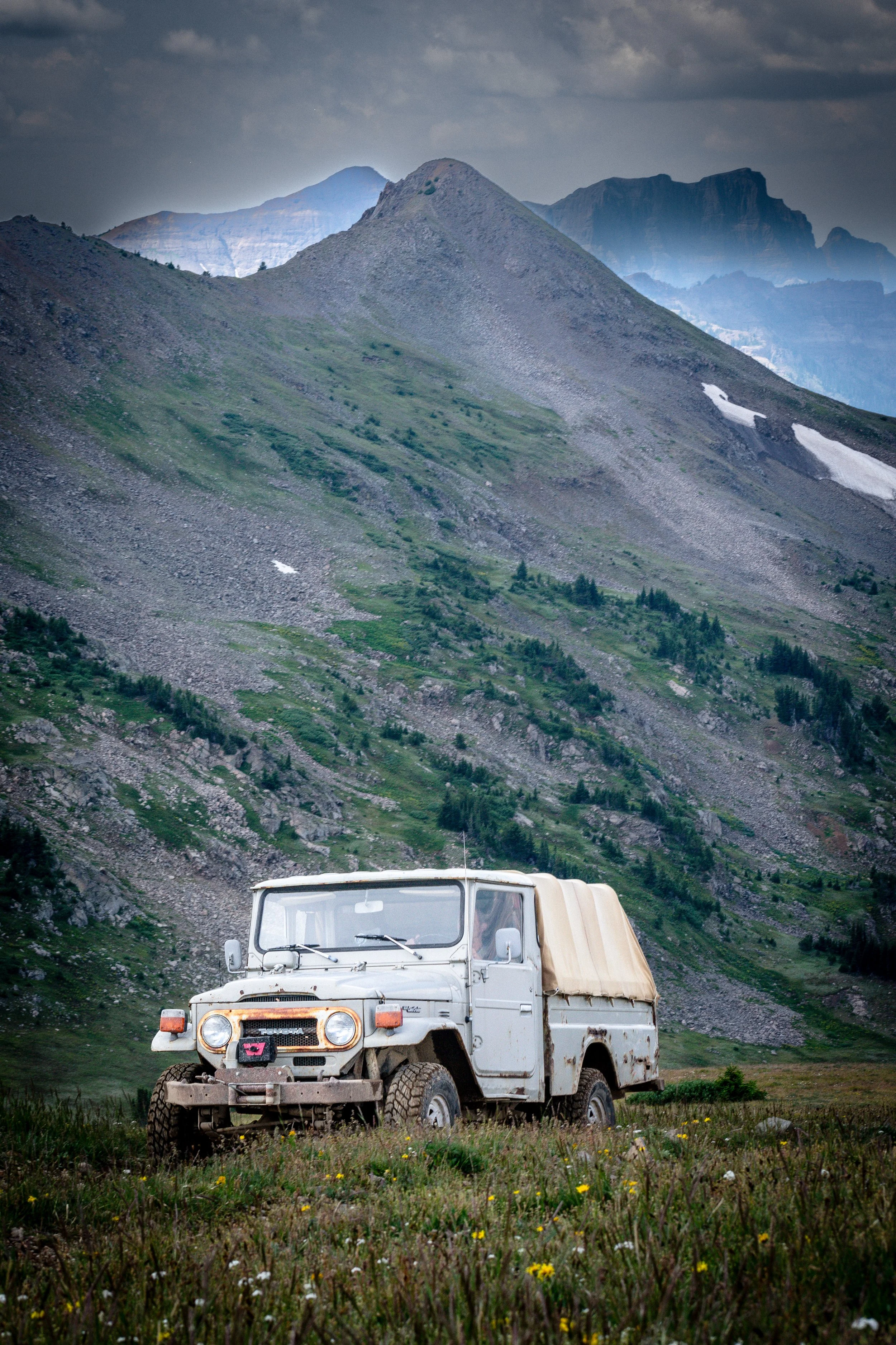 An old, white, rusty pickup truck with a beige cover parked on grassy terrain with wildflowers, set against a backdrop of towering green mountains and cloudy sky.