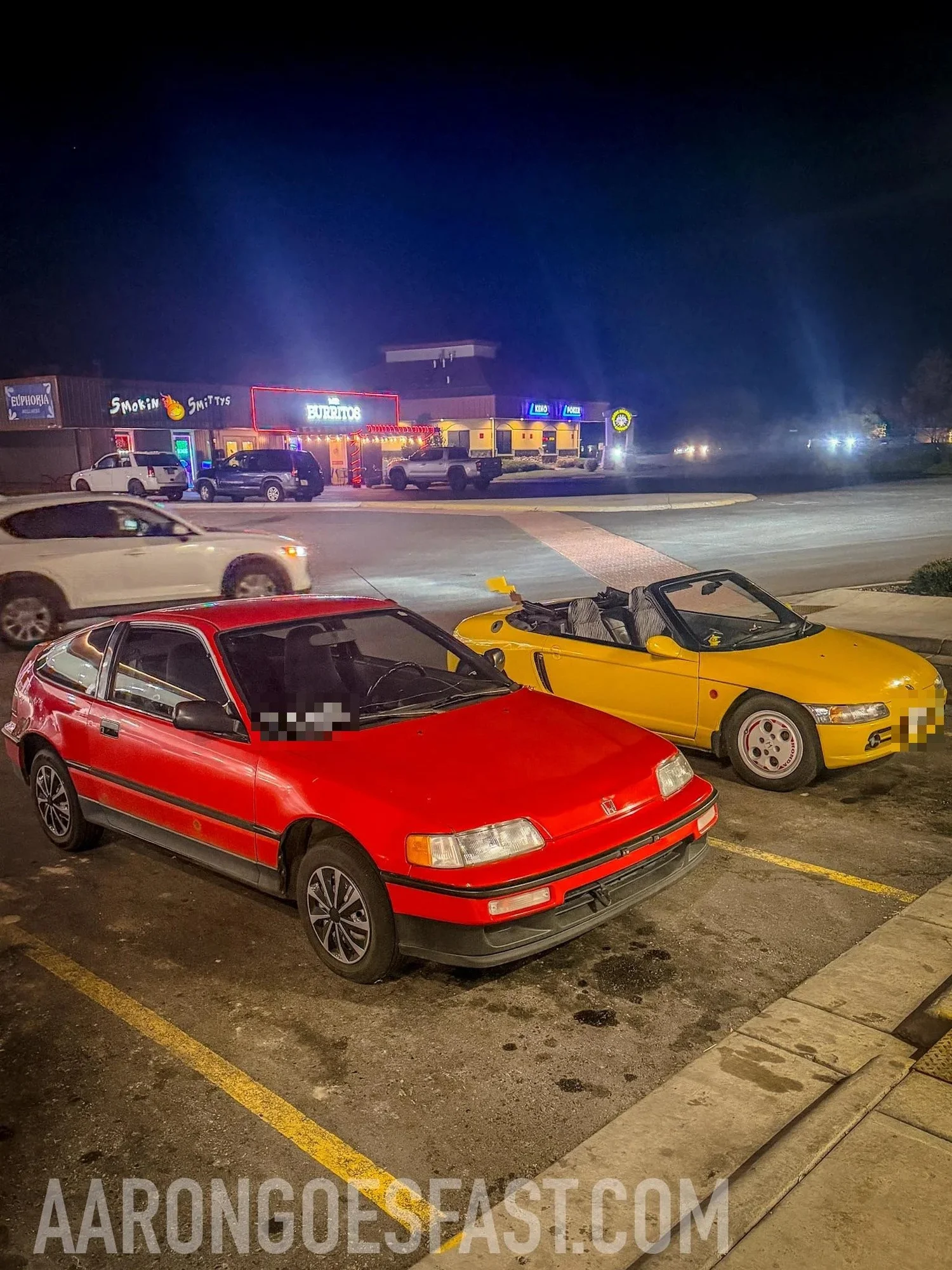 Red second-generation Honda CRX parked next to a yellow Honda Beat convertible at night under neon signs