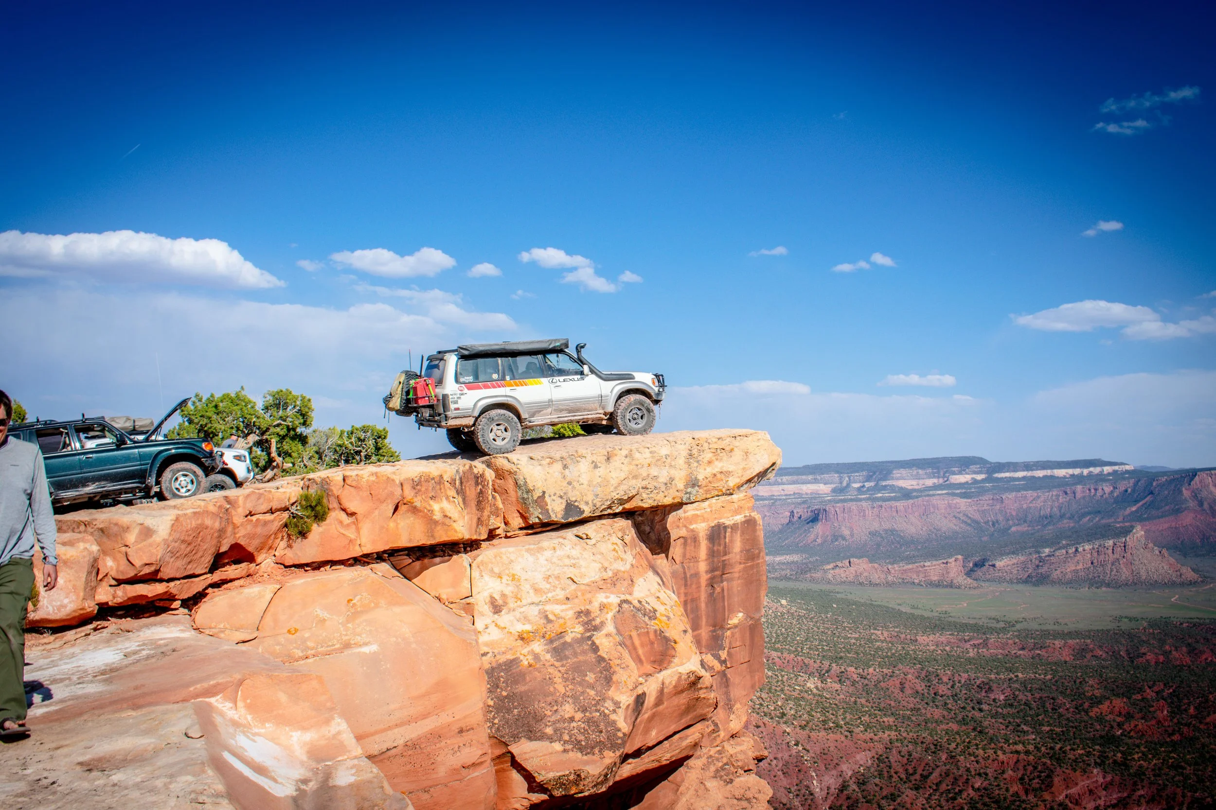 Two off-road vehicles parked on the edge of a rocky cliff at the Grand Canyon, with a person walking nearby and a clear blue sky overhead.