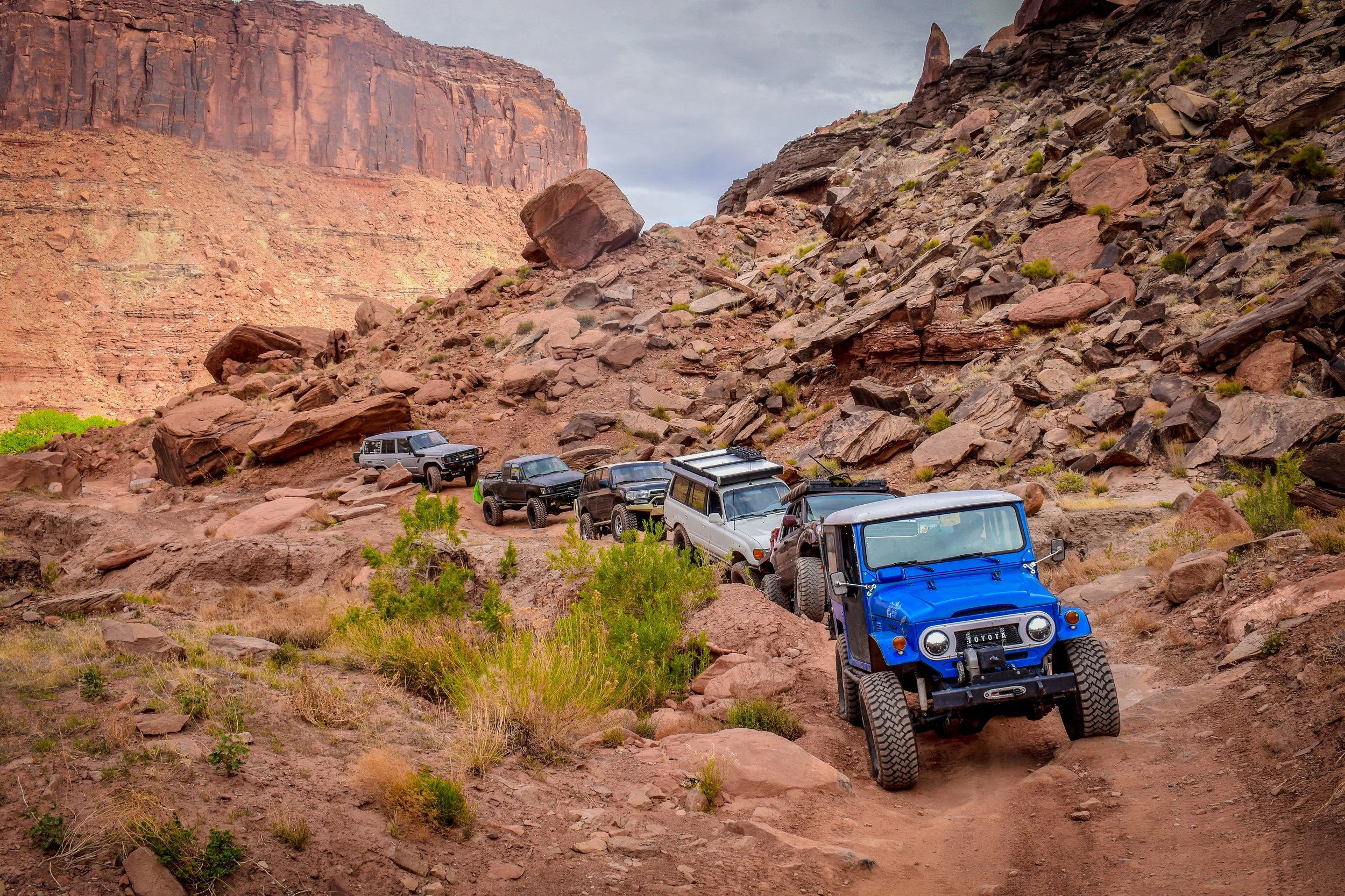 A line of off-road vehicles, including Toyota land cruisers, climbing a rocky trail in a desert canyon with red rock formations.