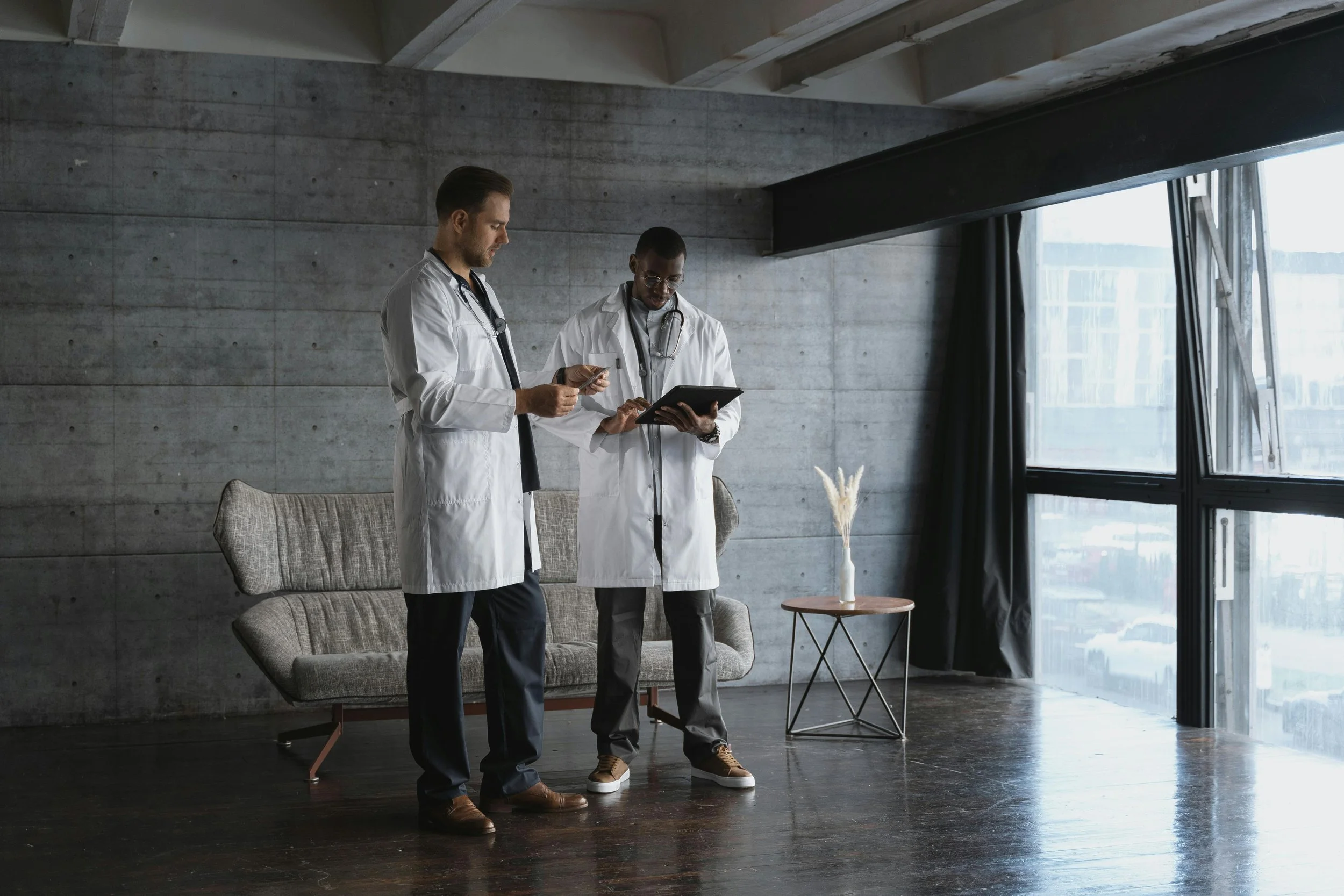 Two male doctors in white lab coats standing inside a modern room with concrete walls, looking at a tablet and a mobile phone.