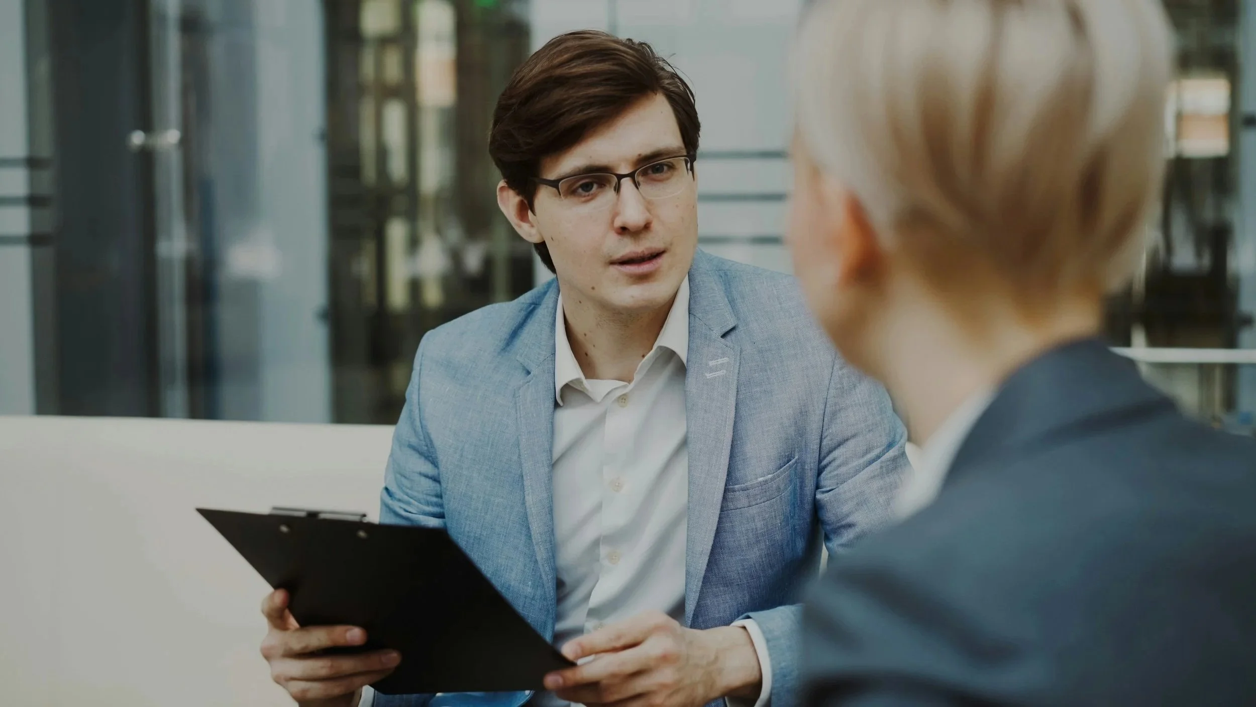 Two men having a business conversation in an office, with one holding a clipboard, engaged in a serious discussion.