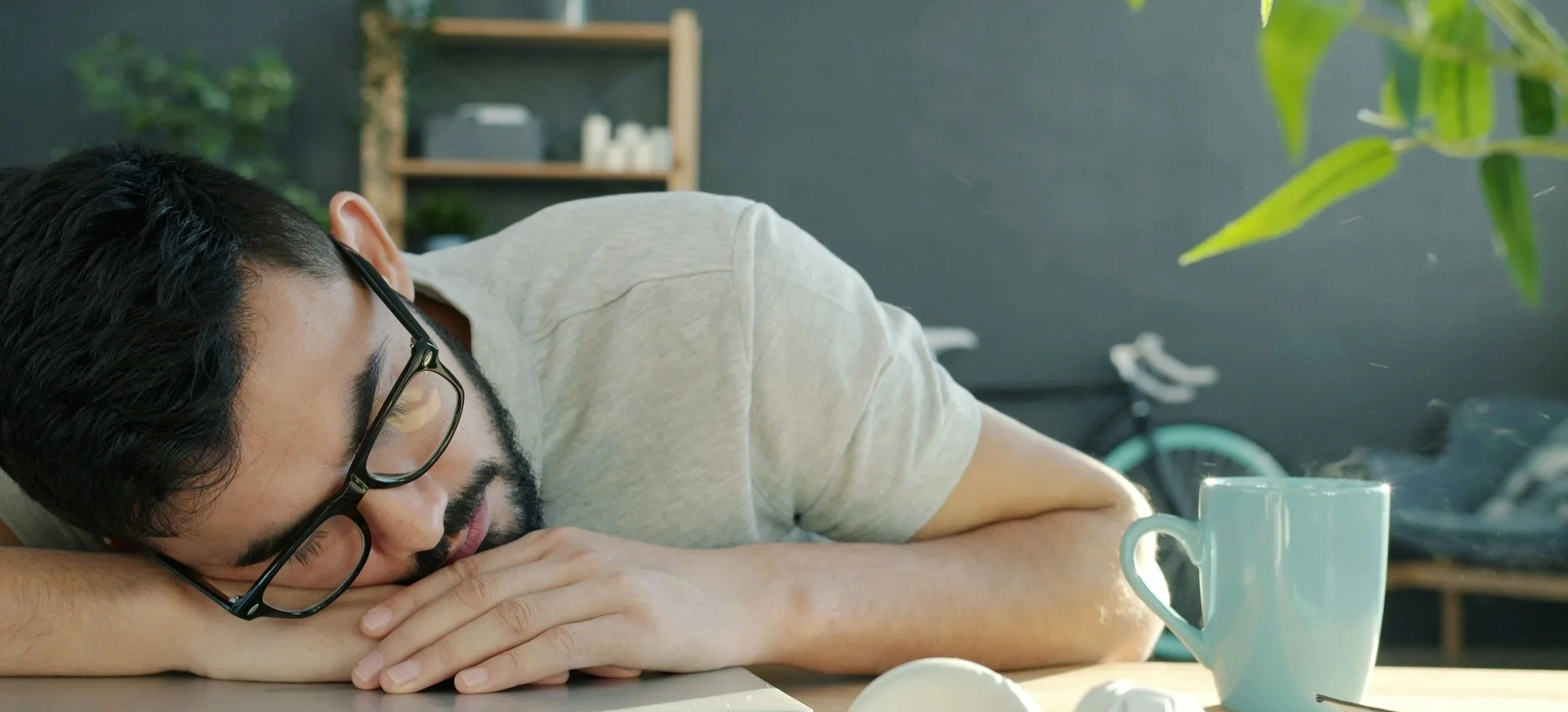 A man with glasses and a beard is sleeping at a table with his head resting on his arm, in a cafe or kitchen setting. There is a white mug on the table and a bicycle in the background.