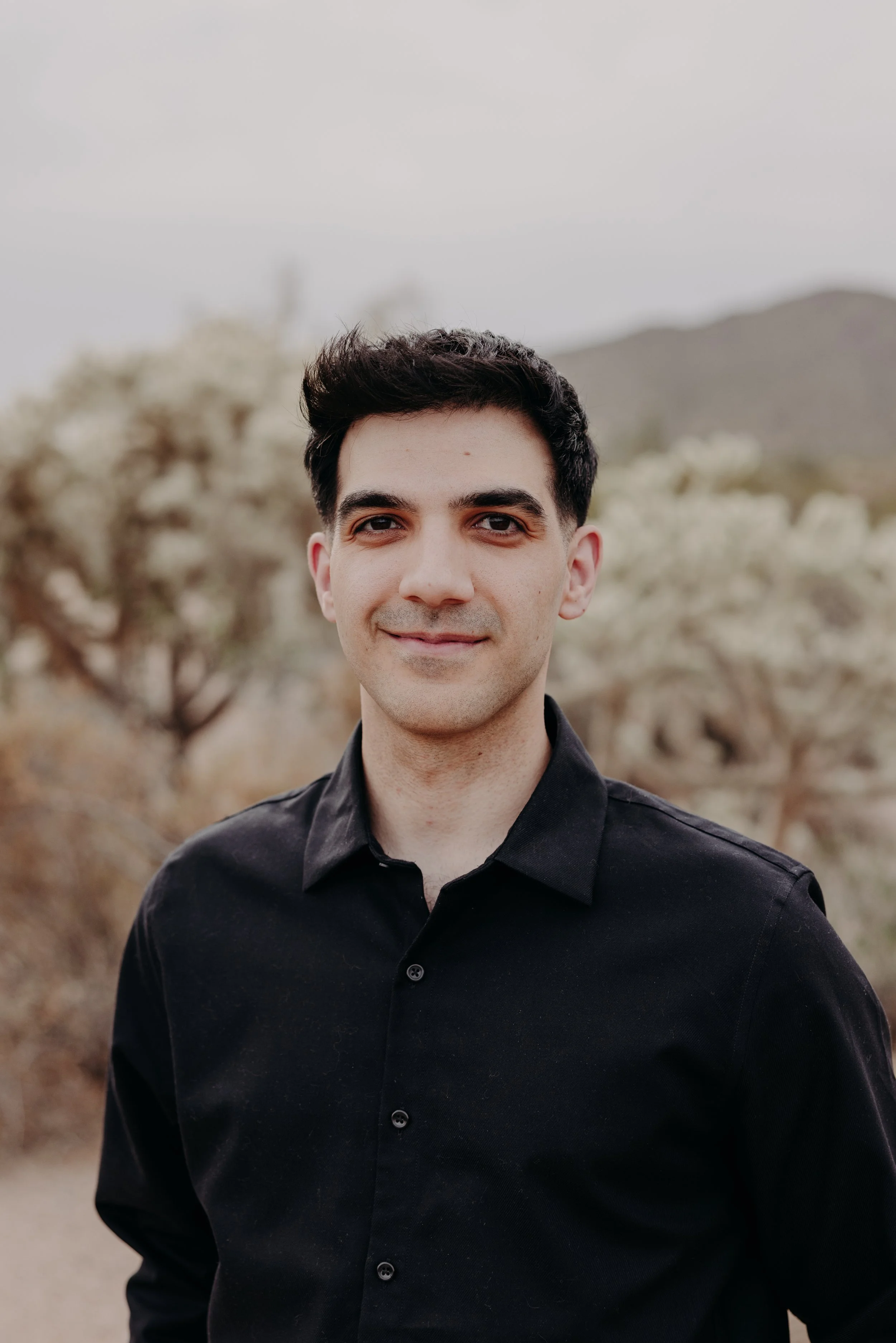 A young man with dark hair and fair skin, wearing a black button-up shirt, standing outdoors with a blurred natural desert landscape background including bushes and mountains.