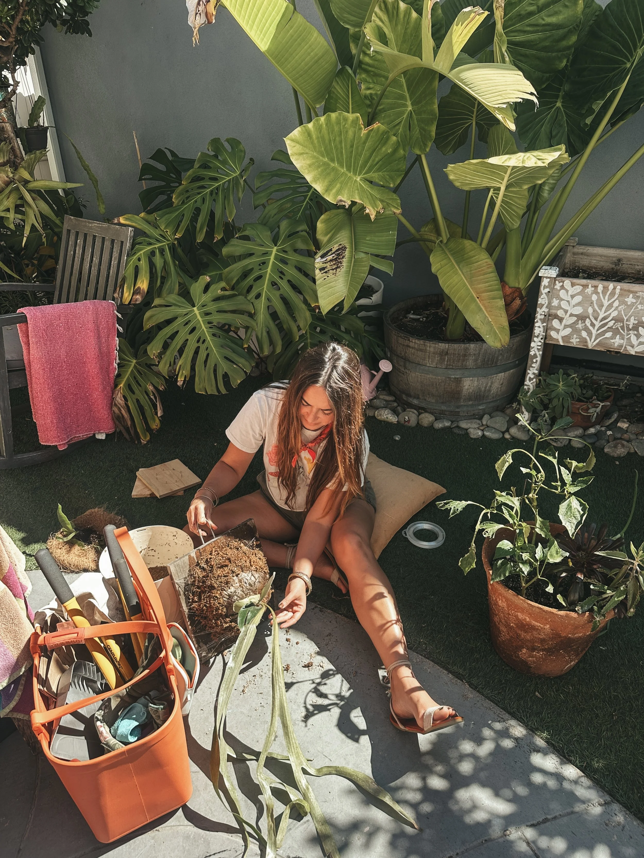 Woman gardening on a patio with large tropical plants, sitting on a cushion, planting soil into a plant pot, with gardening tools and supplies around her.