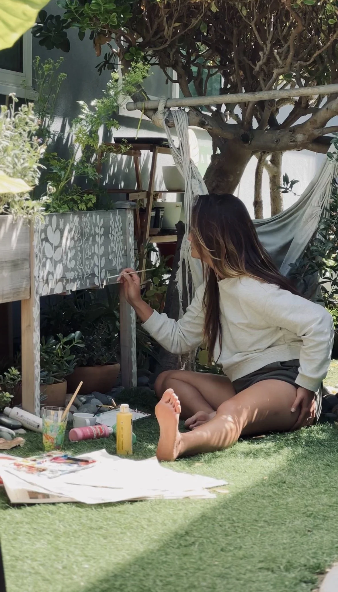 A woman sitting on the grass doing outdoor art painting on a wooden panel, surrounded by plants and garden tools.