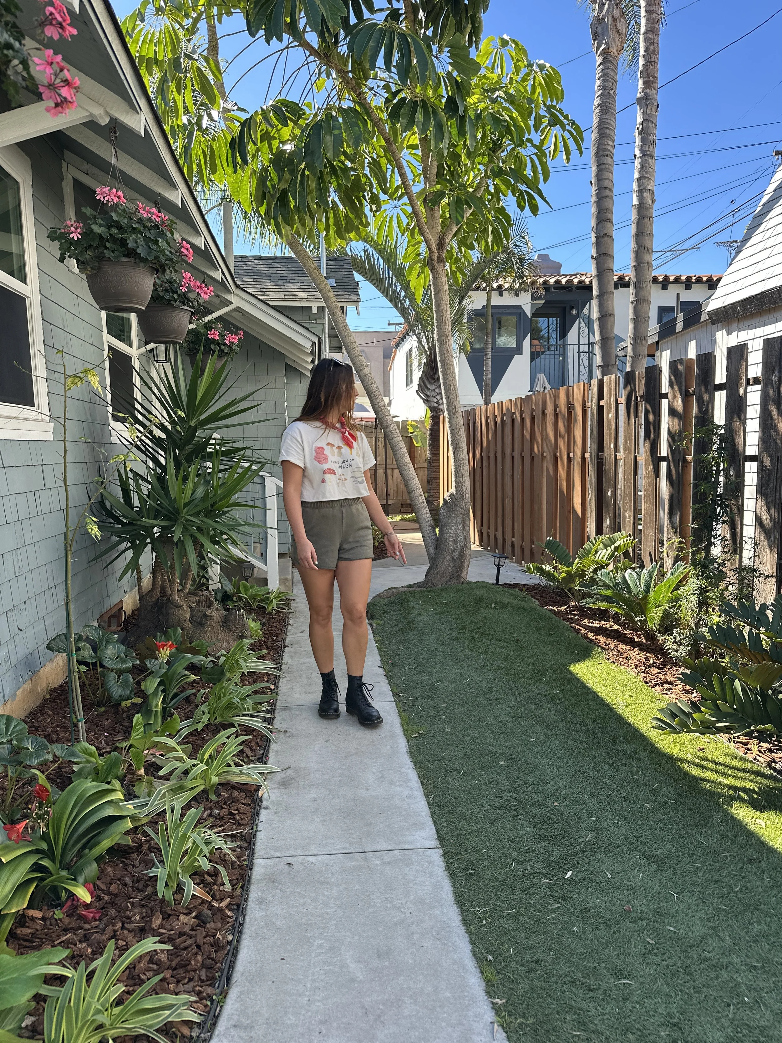 A woman with brown hair wearing a white t-shirt, khaki shorts, and black boots walking on a concrete sidewalk in a garden yard with various plants, trees, and hanging flower pots.