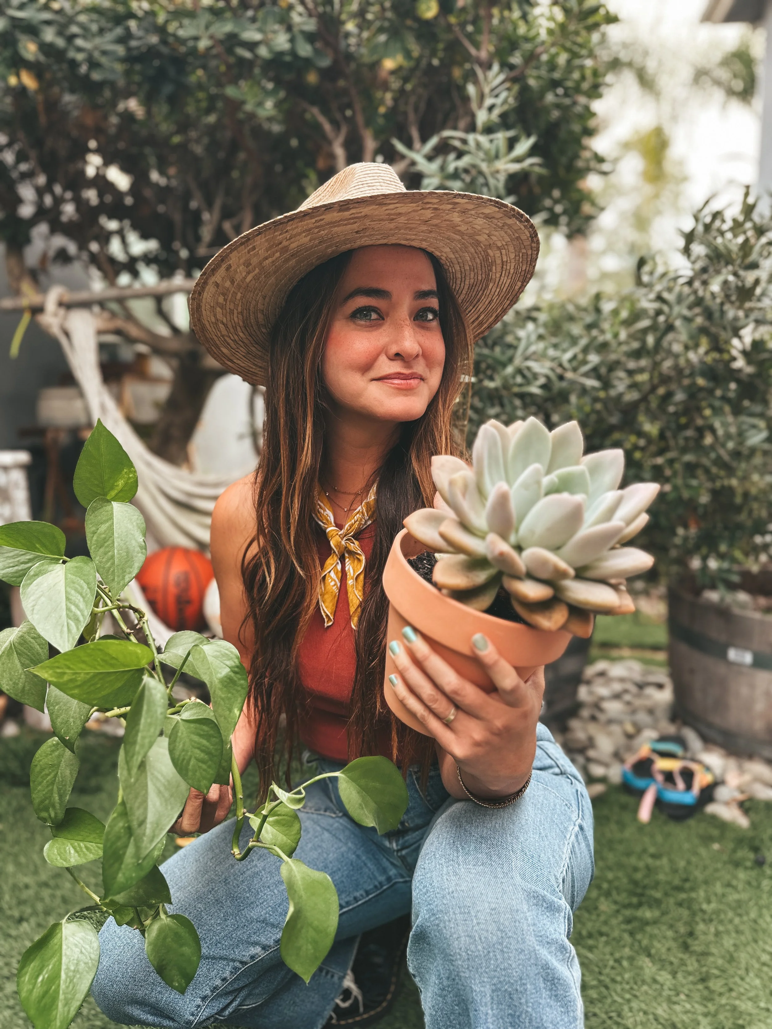Young woman wearing a wide-brimmed straw hat, holding a potted succulent plant, sitting outdoors in a garden with plants and trees.