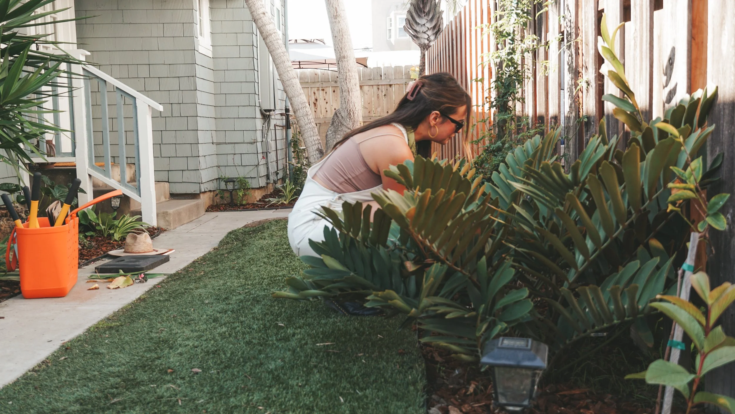 A woman kneeling in a backyard garden, tending to green shrub-like plants along a wooden fence. She is wearing sunglasses, a sleeveless top, and white pants, with gardening tools and supplies nearby.