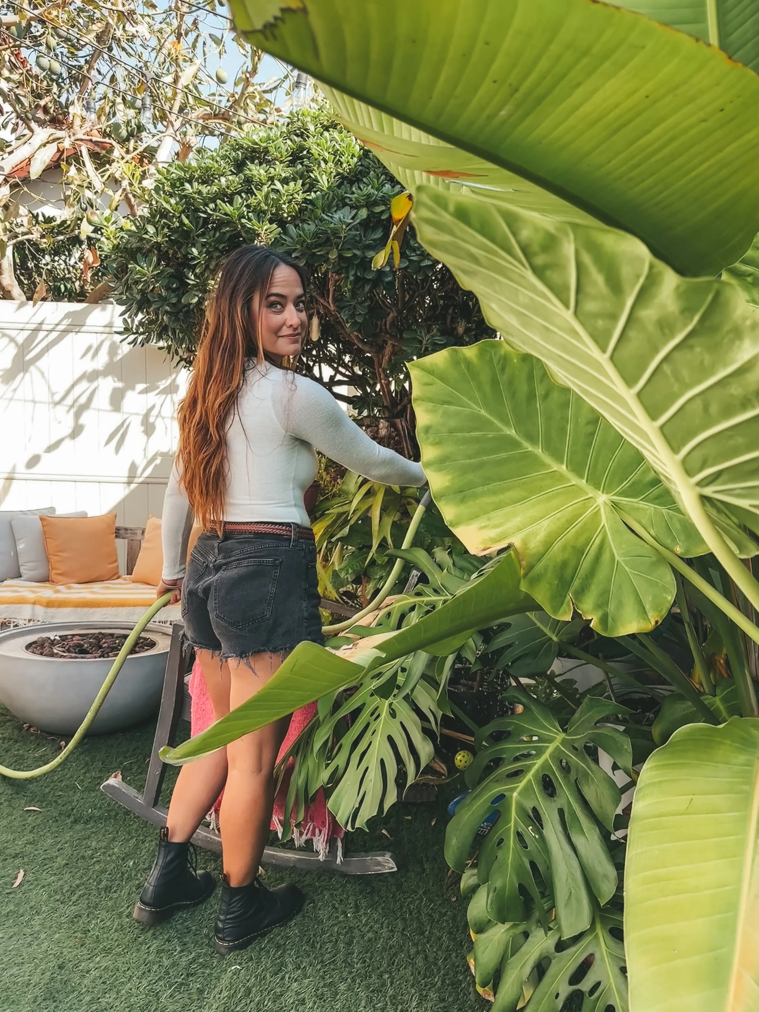 A woman with long brown hair wearing a white long-sleeve top, black denim shorts, and black boots standing among large green tropical leaves in a backyard garden.