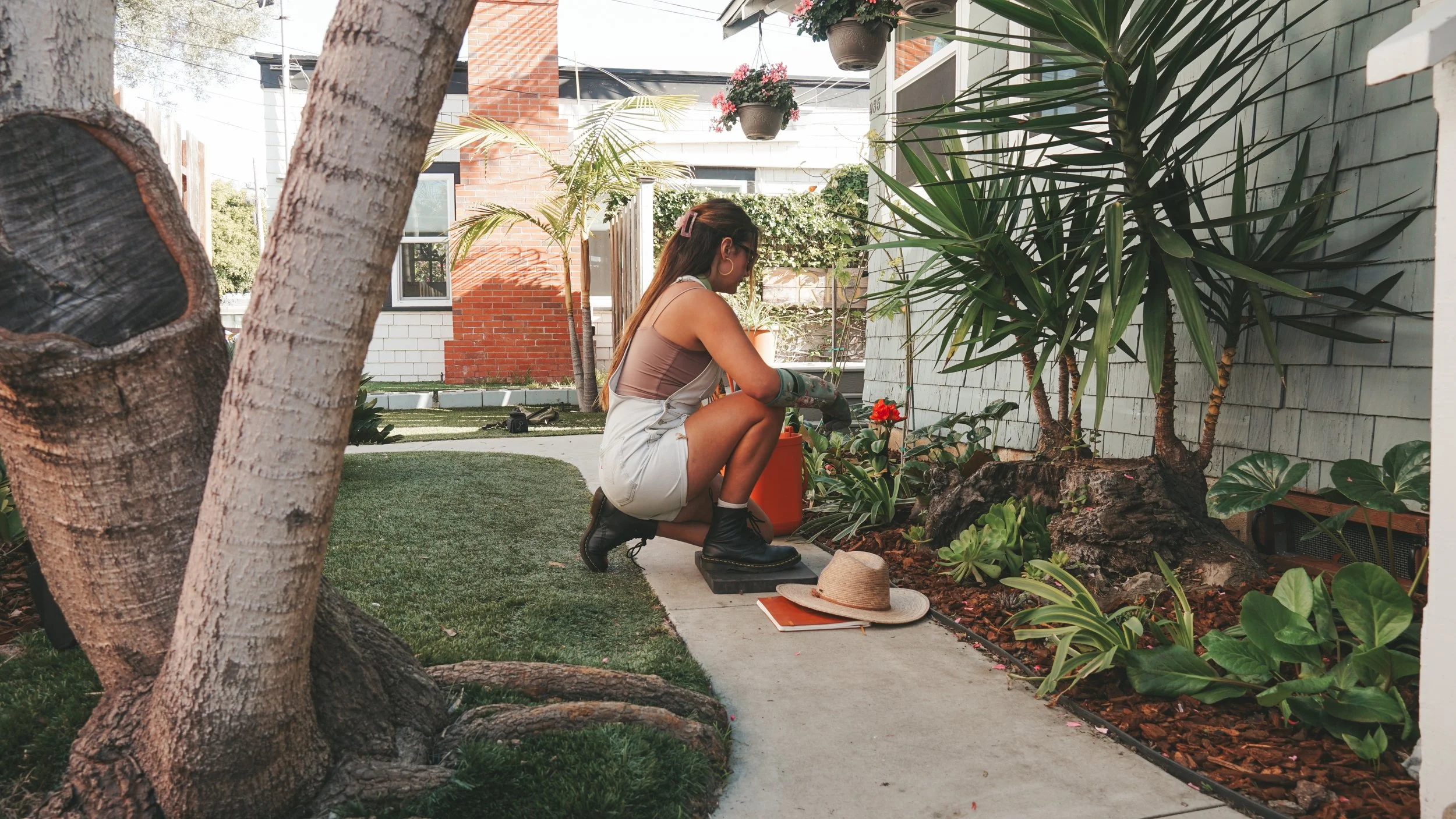 A woman gardening in a backyard with plants, trees, and a brick and white house in the background.
