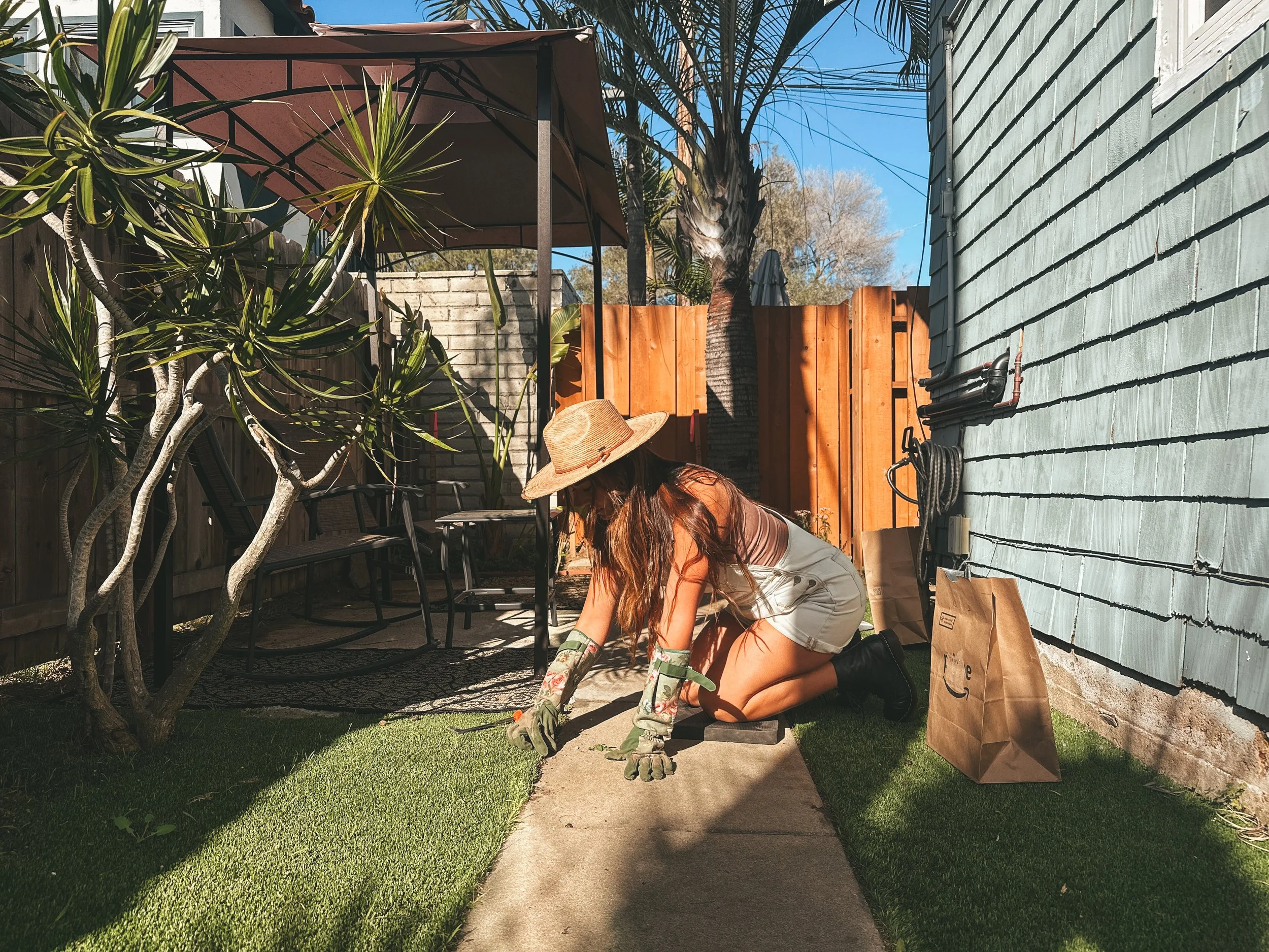 A woman wearing a large straw hat, gardening gloves, a tank top, and shorts, kneeling on a concrete pathway while planting or weeding in a backyard garden. The yard has a wooden fence, a blue house wall, and various plants and outdoor furniture.