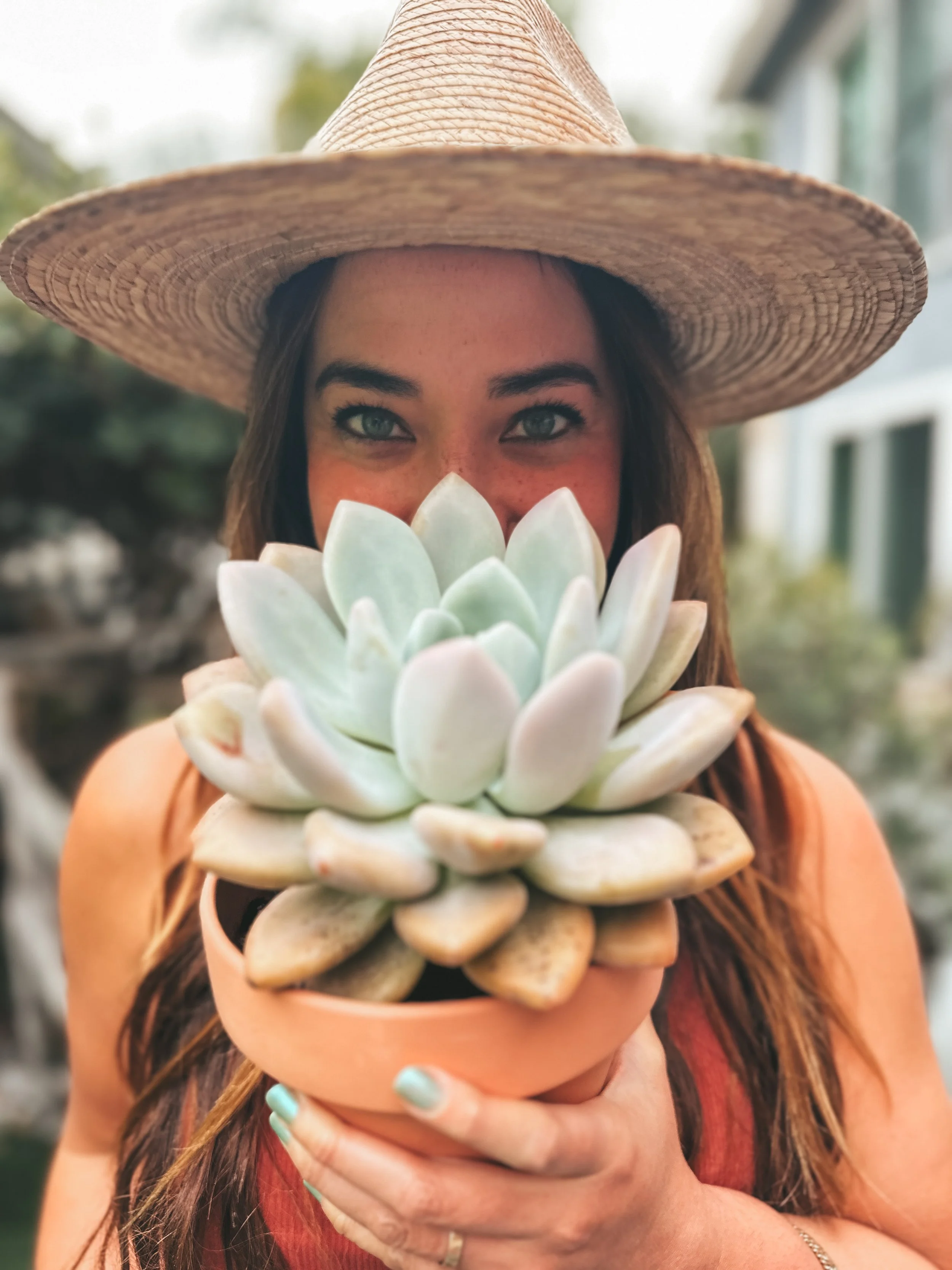 A woman with blue eyes wearing a wide-brimmed straw hat and red tank top, holding a potted succulent plant in front of her face, with a blurred outdoor background.