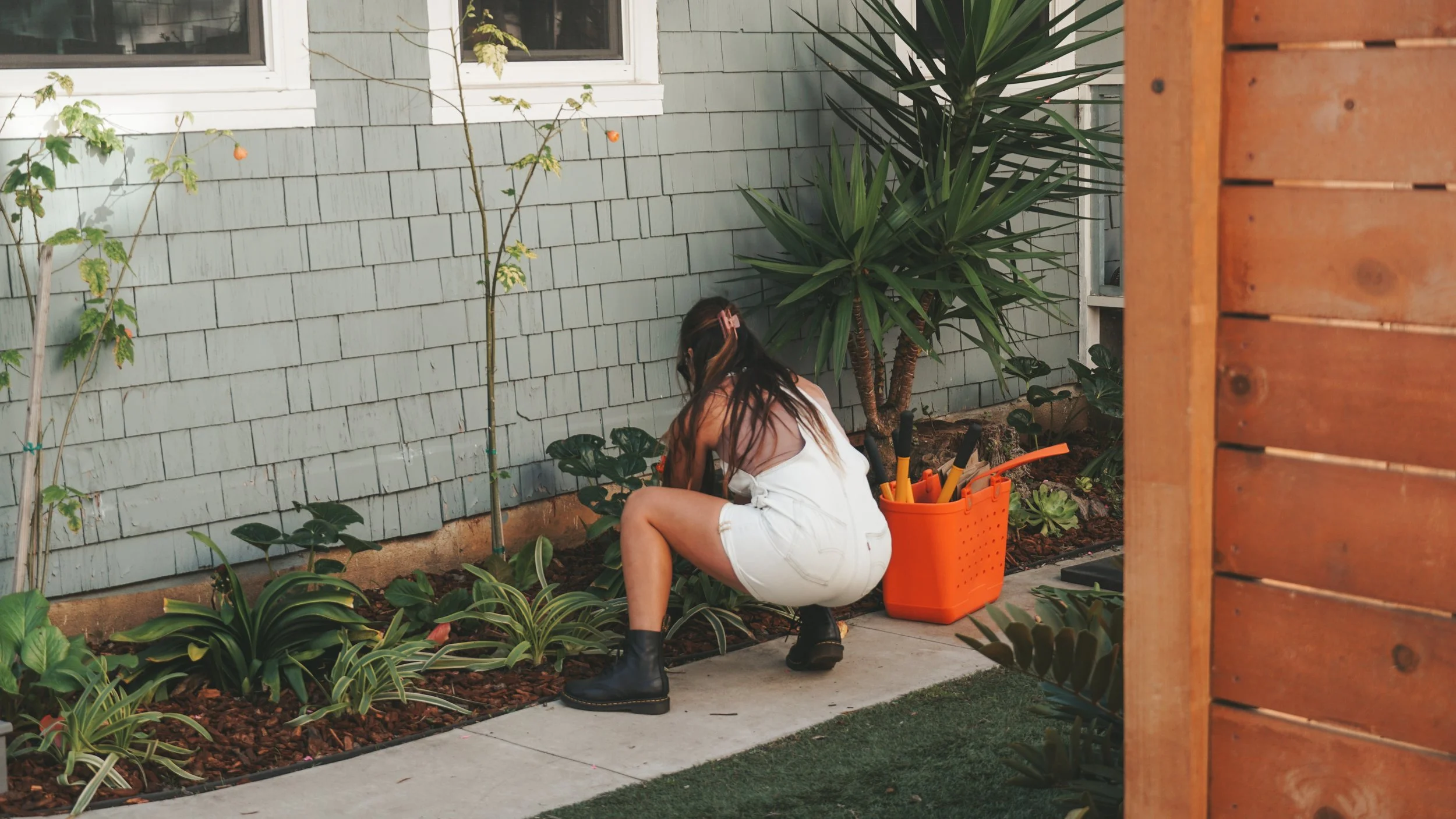 A woman with long dark hair wearing a white top, white shorts, and black boots kneeling in a garden bed, planting or tending to plants next to a purple house wall. An orange container with gardening tools is beside her.