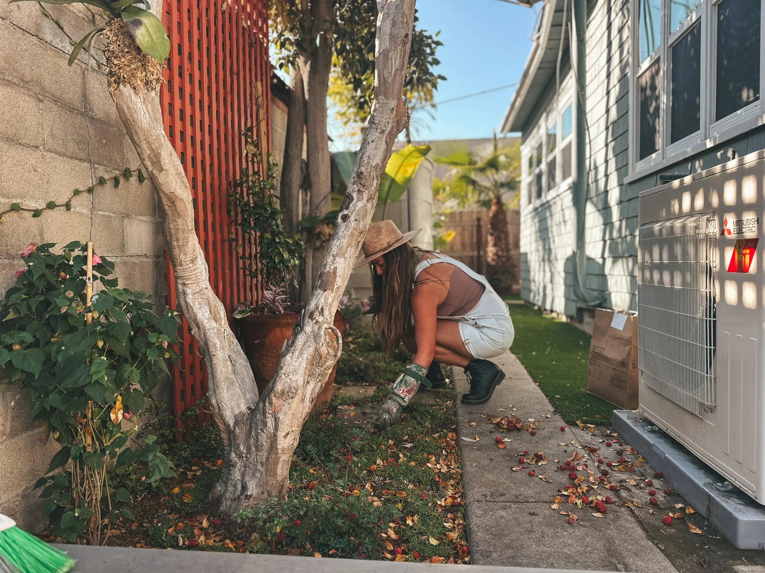 A woman wearing a hat, gloves, and boots is gardening in a backyard, crouched near a tree and planting or tending to flowers and plants along a garden bed.
