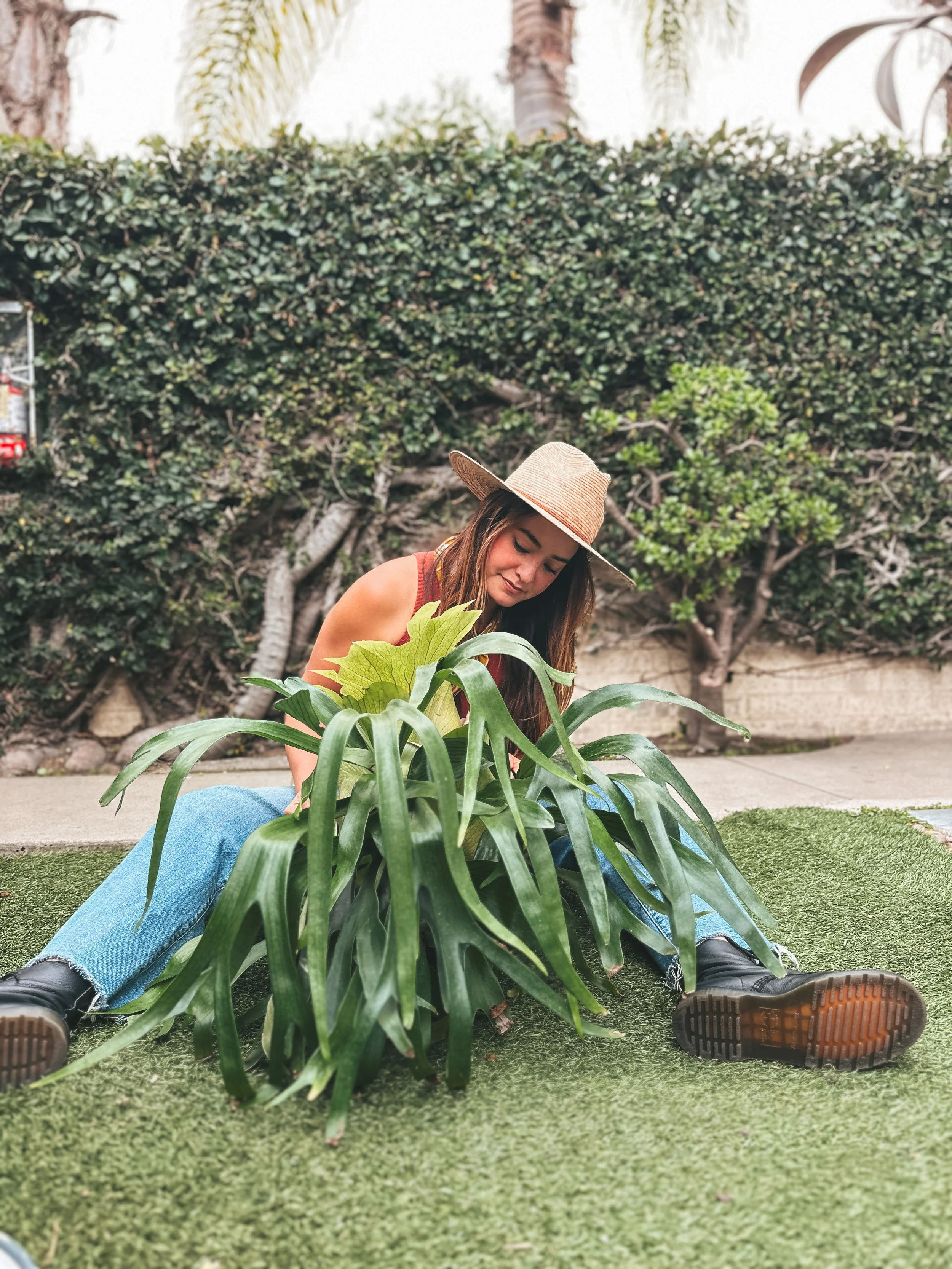 A woman wearing a straw hat and black boots sitting on the ground beside a large leafy green plant in an outdoor garden or yard.