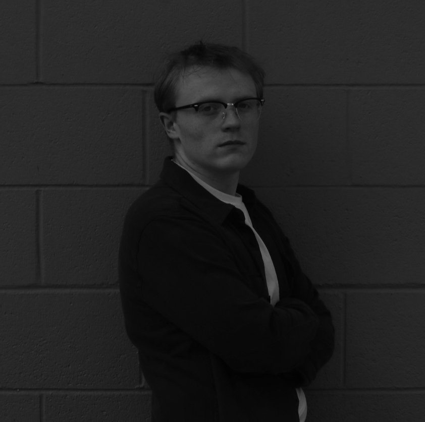 A young man wearing glasses and a black jacket standing against a brick wall, with his arms crossed and looking at the camera.