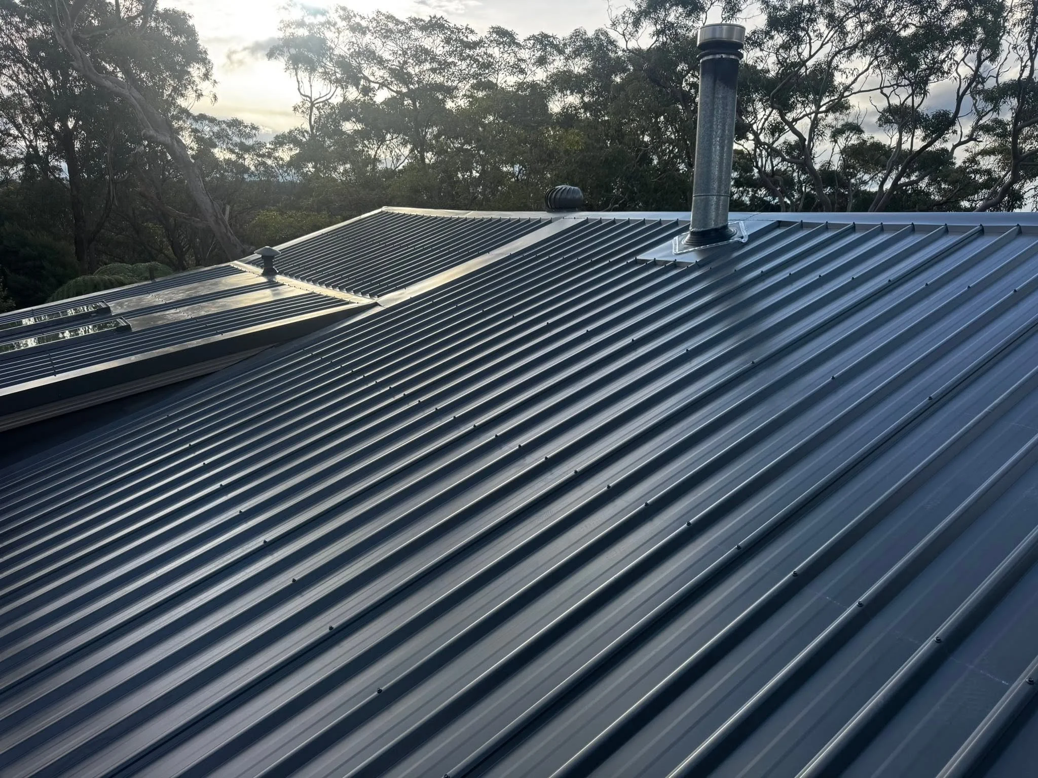 Close-up of a metal roof with three vents and a chimney, surrounded by trees with cloudy sky in the background.