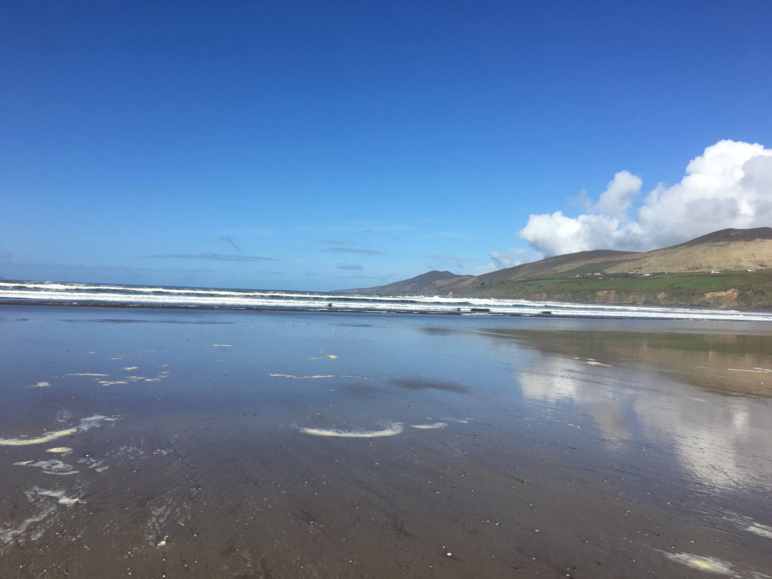 Photo of low tide at Dingle Bay from Castlemain Harbor near Inch, Ireland. Hills rise high across the bay under fluffy clouds and blue skies, their image mirrored on the wet sand.