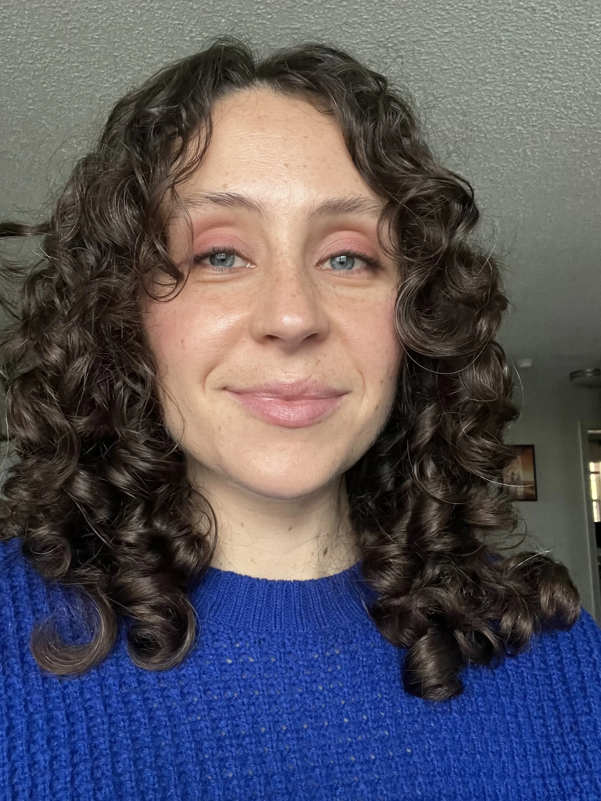 Photo of Lauren smiling in a blue knit sweater. She has brown curly hair, blue eyes, and white skin.