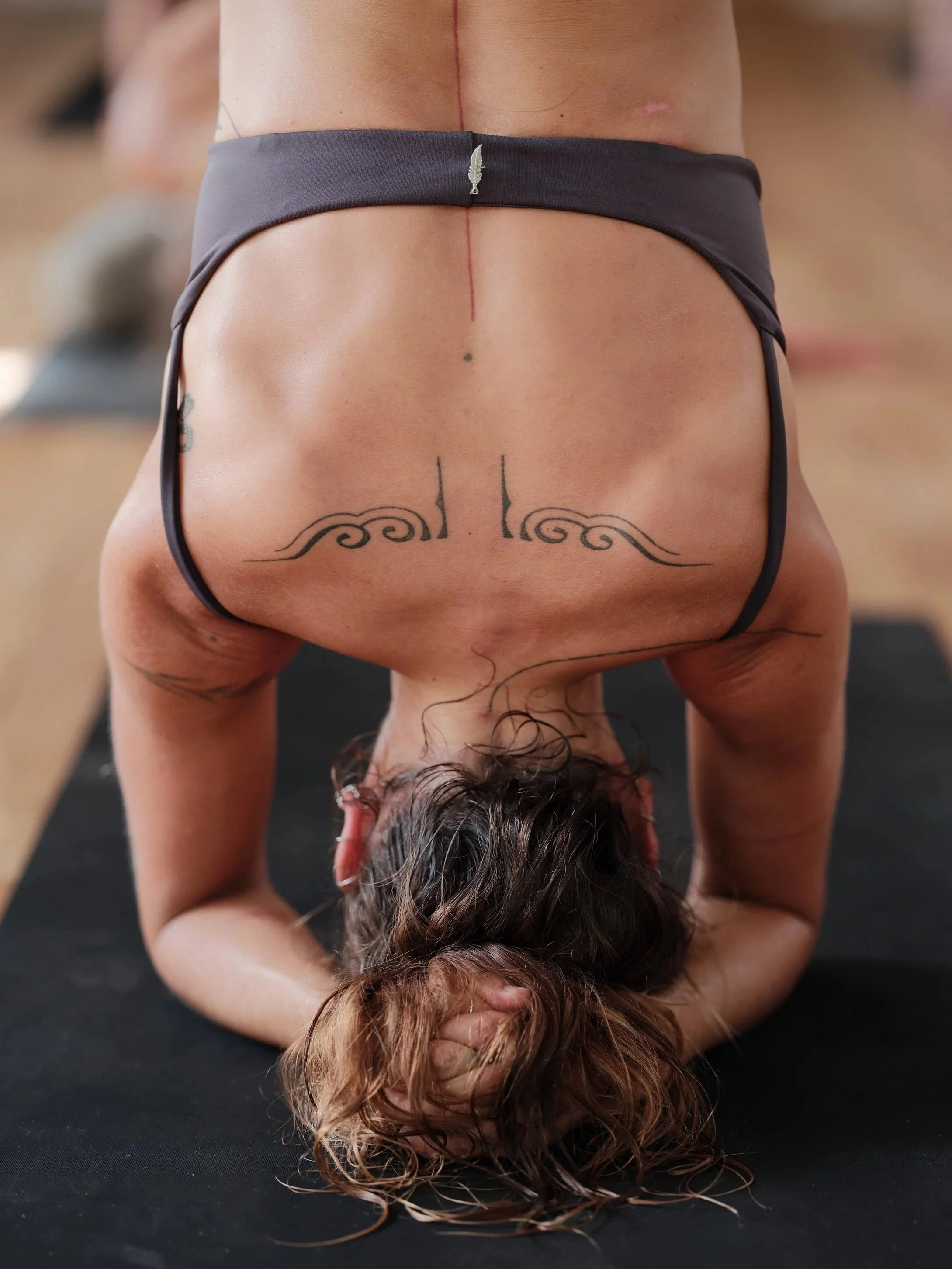 A person performing a forward bend yoga pose, with their head and hands on the ground, wearing a sports bra, and tattoos visible on their back and arms.