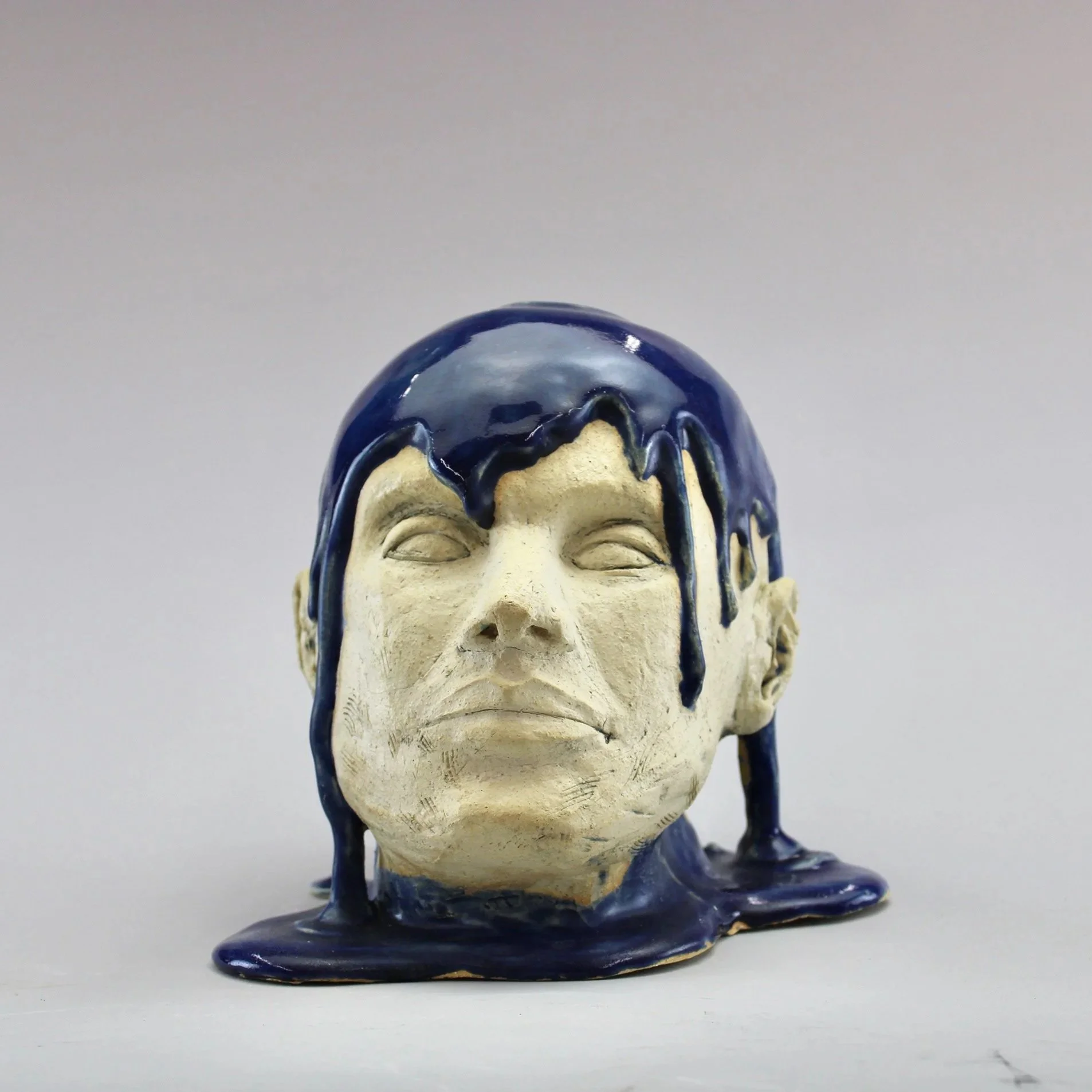 Sculpture of a human face with dark blue, glossy liquid dripping down from skull against a plain light background by Anna Ford.