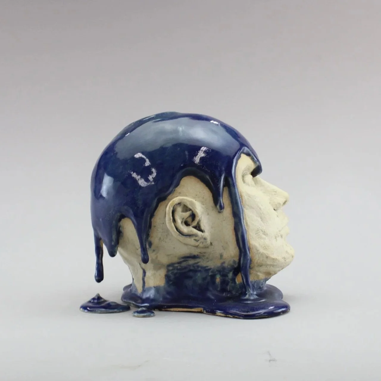 Sculpture of a human face with dark blue, glossy liquid dripping down from skull against a plain light background by Anna Ford.