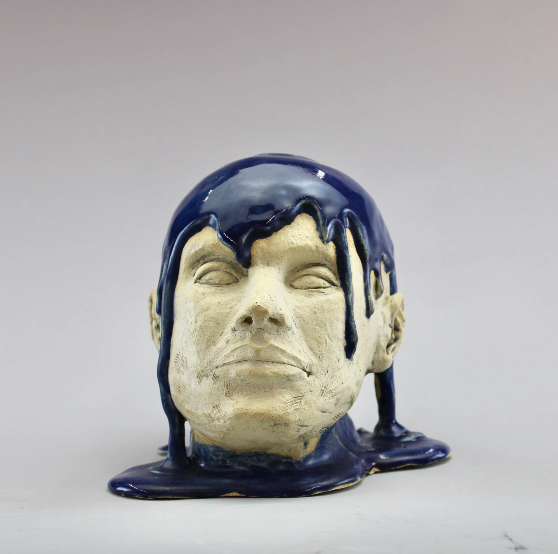 Sculpture of a human face with dark blue, glossy liquid dripping down from skull against a plain light background by Anna Ford.
