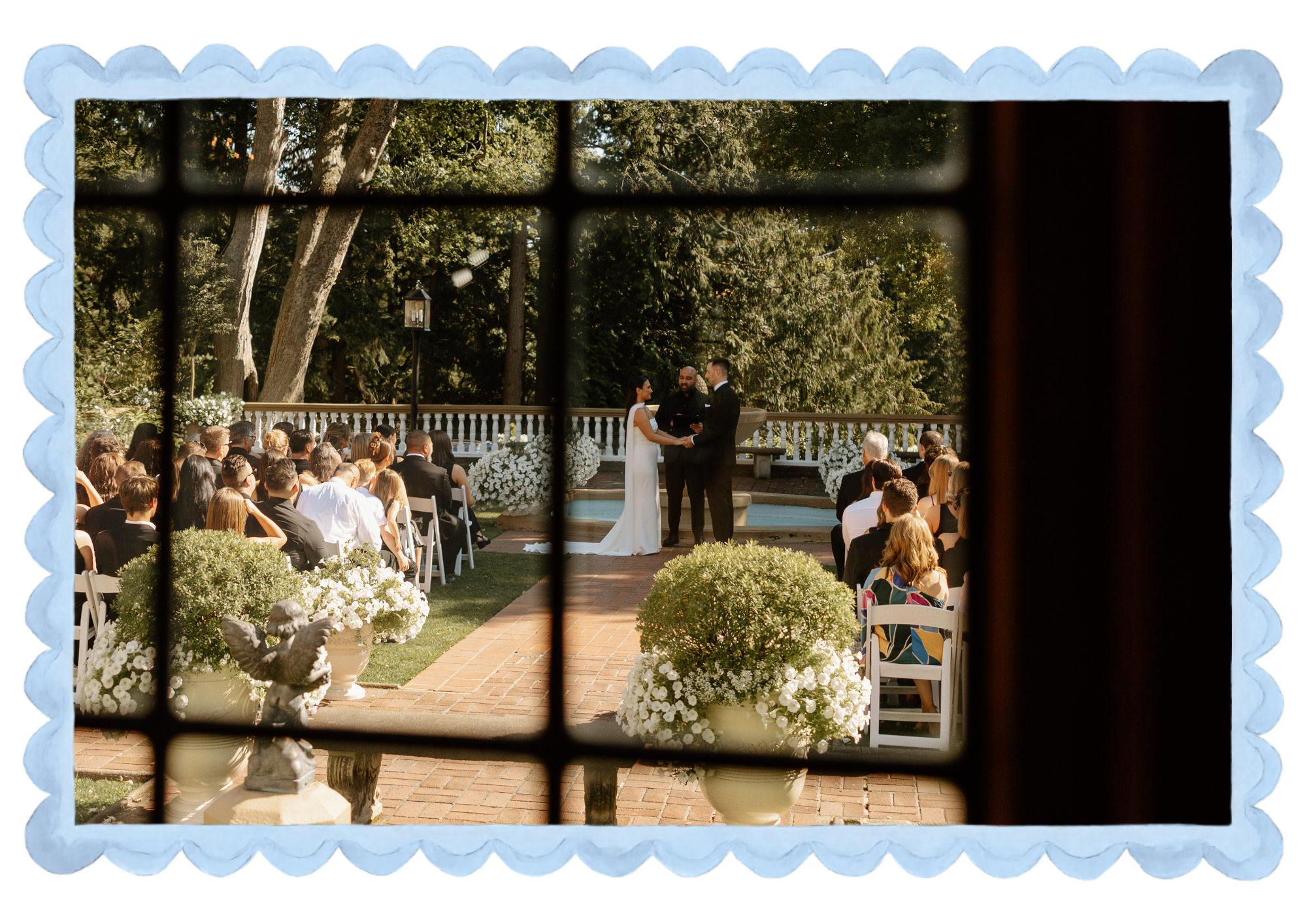 An outdoor wedding ceremony viewed through a black window frame with a decorative light-blue border. The couple is standing at the altar holding hands, with an officiant between them. Guests are seated on white chairs, and the scene has lush greenery and trees in the background.