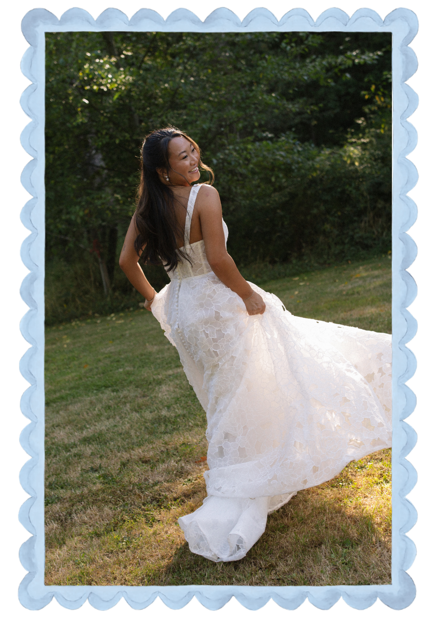 A woman in a wedding dress outdoors on grass, smiling, with trees in the background.