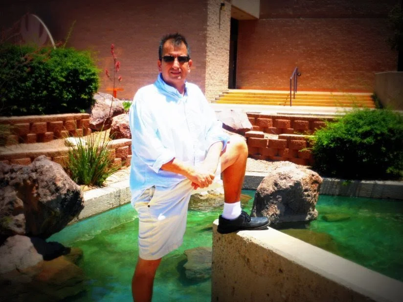 Man standing on a stone ledge with one foot raised, in a pond area with rocks and greenery, outside brick building in the background.