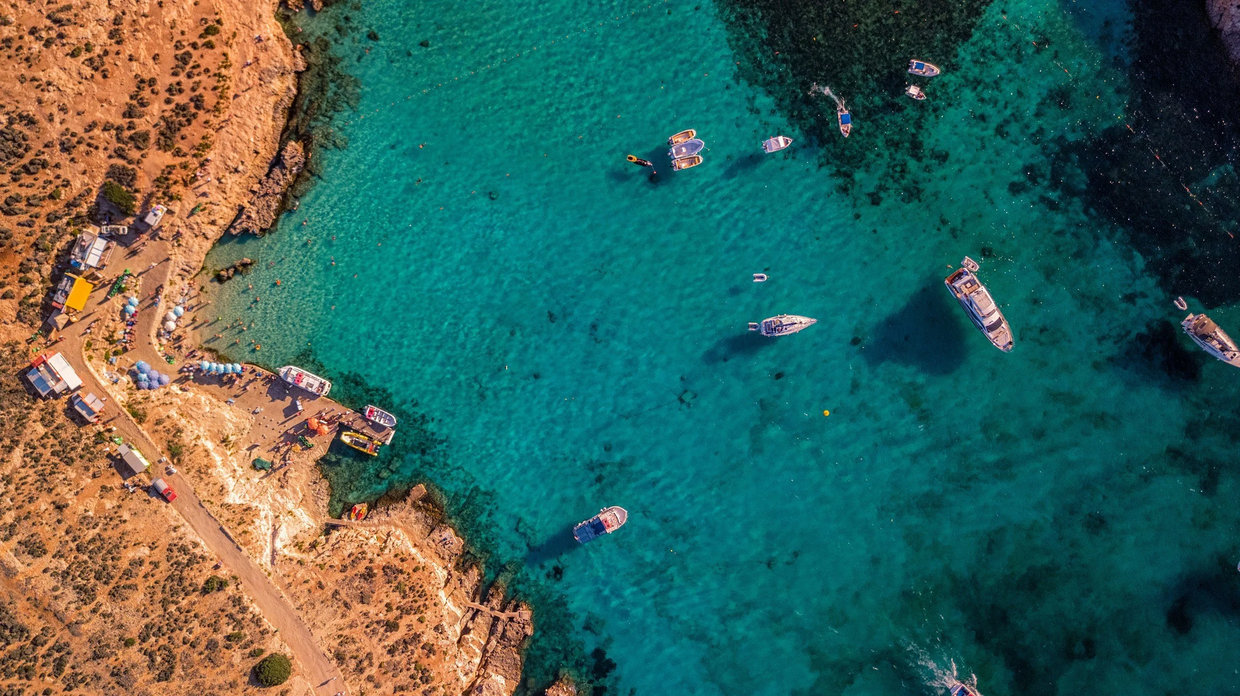 Aerial view of a coastline with clear blue water, small boats anchored near the shore, and a rocky beach area with tents and people.