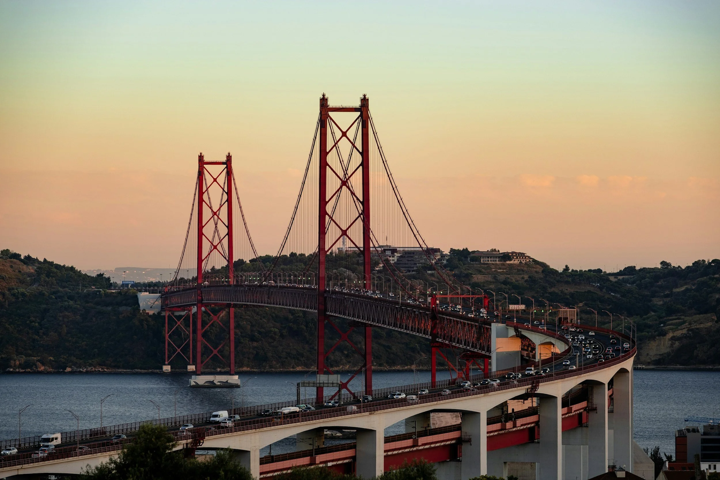 A large orange suspension bridge spans over water, with many cars driving across. Hills and buildings are visible in the background under a sky with soft pastel colors.