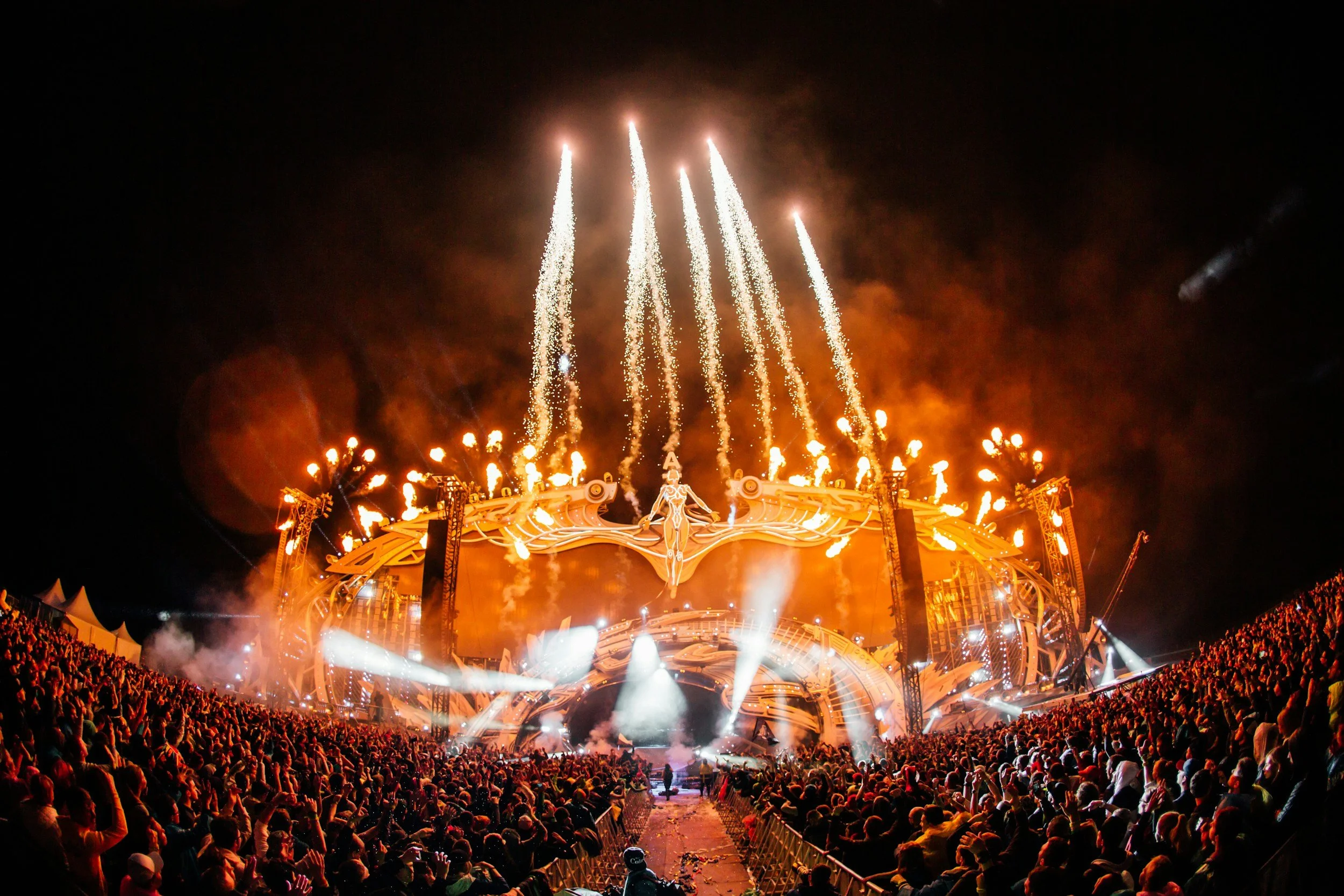 A large crowd gathered at a nighttime music festival with fireworks and bright lights illuminating the stage.