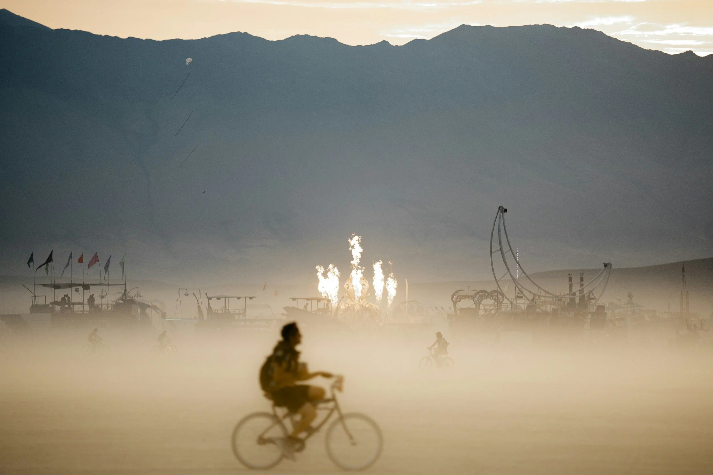 A person riding a bicycle in a desert at sunset with mountains in the background. There is a large outdoor event with flags and amusement rides, and fireworks lighting up the sky.