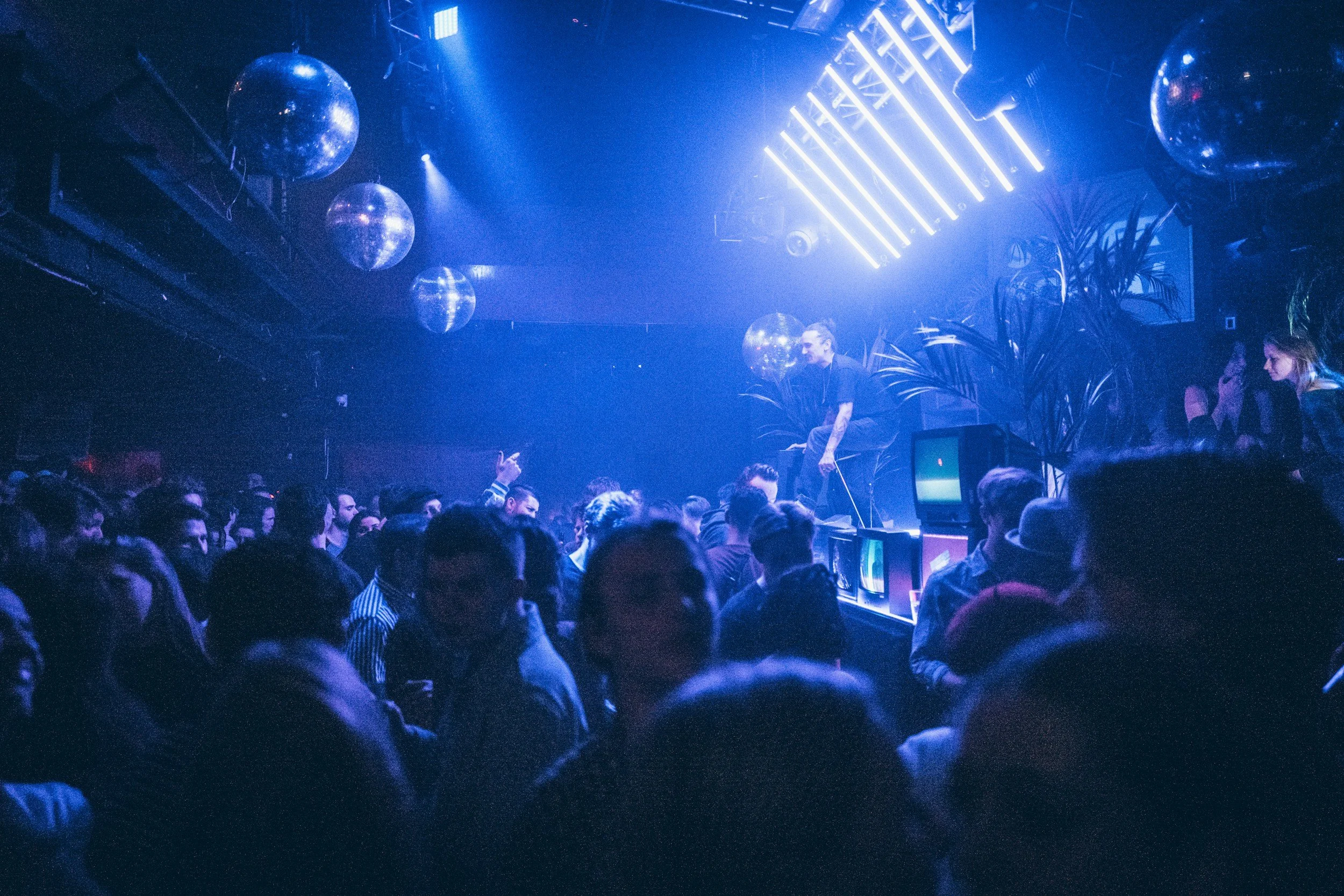Crowd of people at a nightclub dance floor with DJ performing on stage, illuminated by blue and purple lights, large disco balls, and hanging light fixtures.
