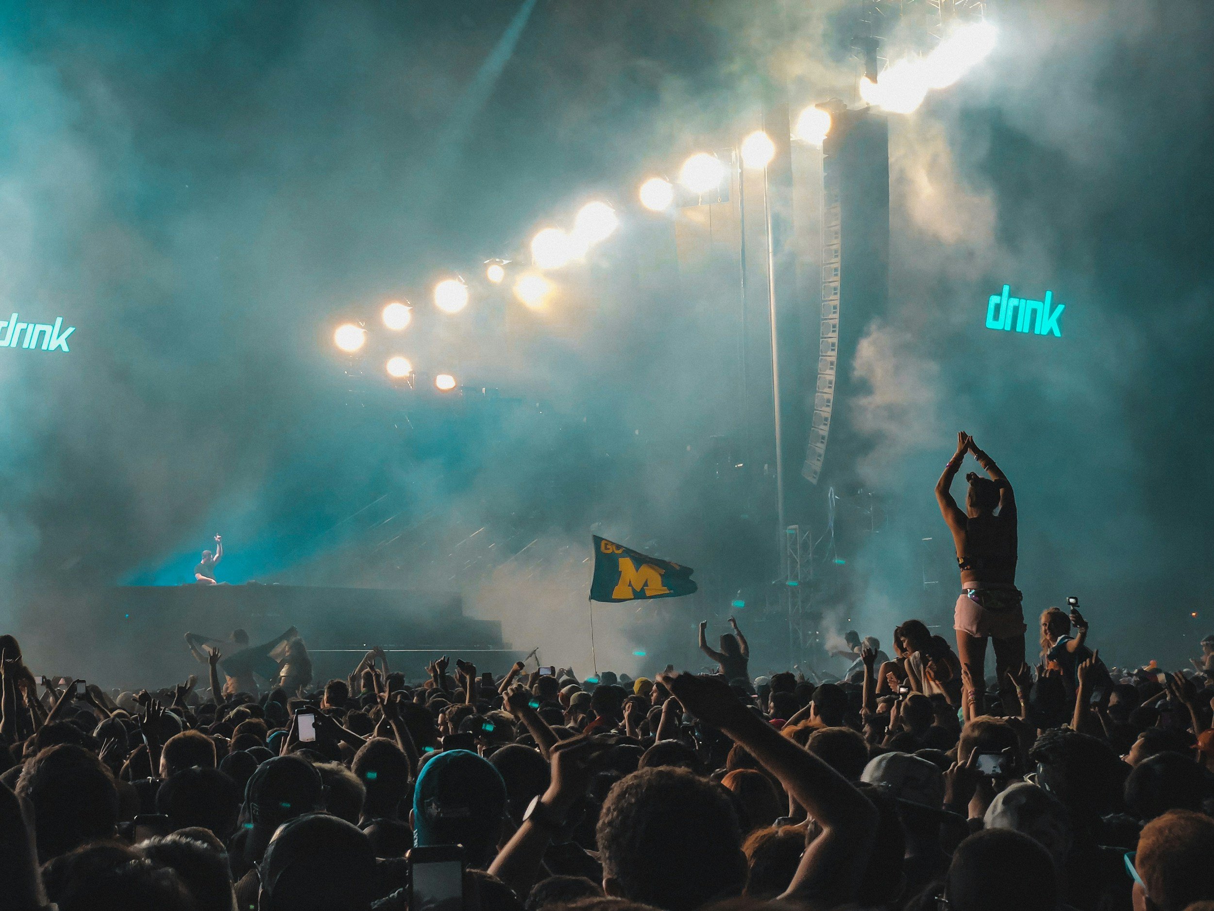 A concert scene with a large crowd and stage lighting, people dancing and taking photos, with a person on stage and a flag with the University of Michigan logo.