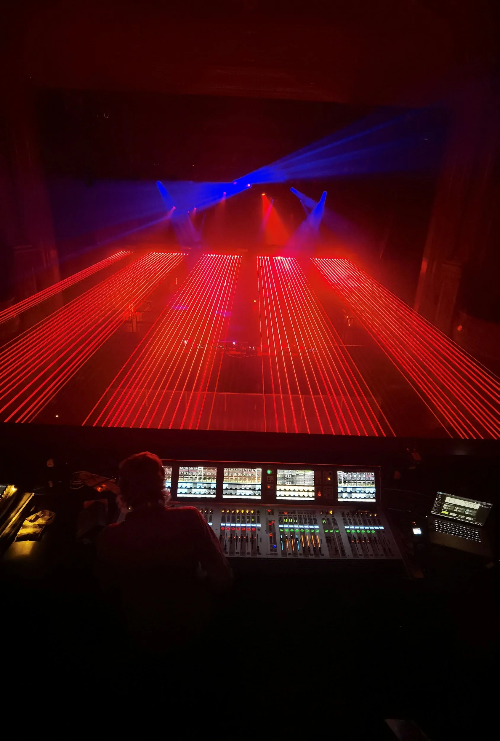 A stage with red and blue laser lights shining into a dark venue, with a DJ's mixing desk and a person operating it visible in the foreground.