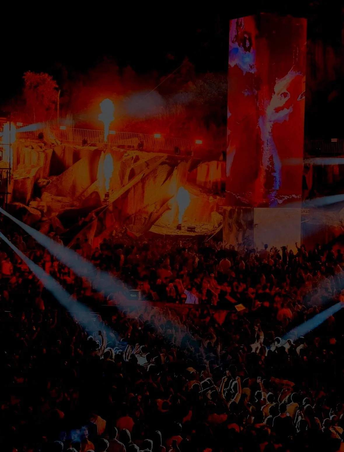 A large crowd at a concert or festival at night with bright stage lights, fire effects, and a large screen displaying visuals.