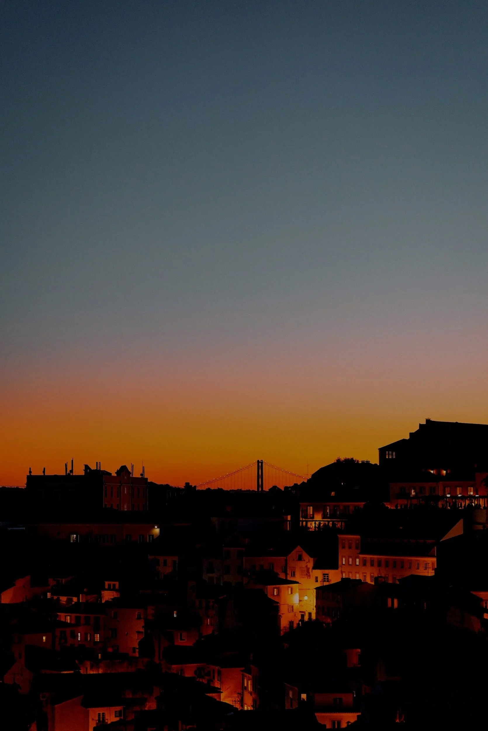 Cityscape at sunset with dark silhouettes of buildings and a bridge, and a colorful sky transitioning from orange to dark blue.