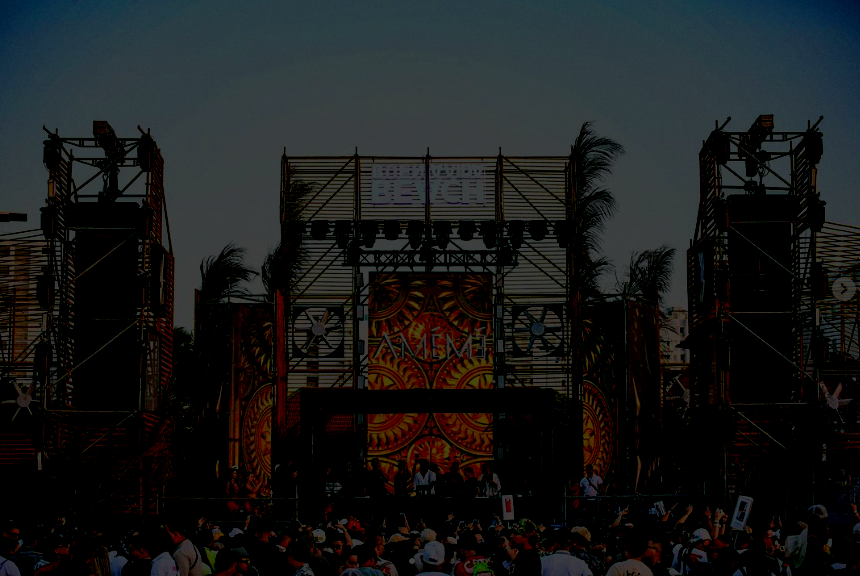 Concert stage with pyrotechnics and a crowd of festival attendees at sunset or dusk.