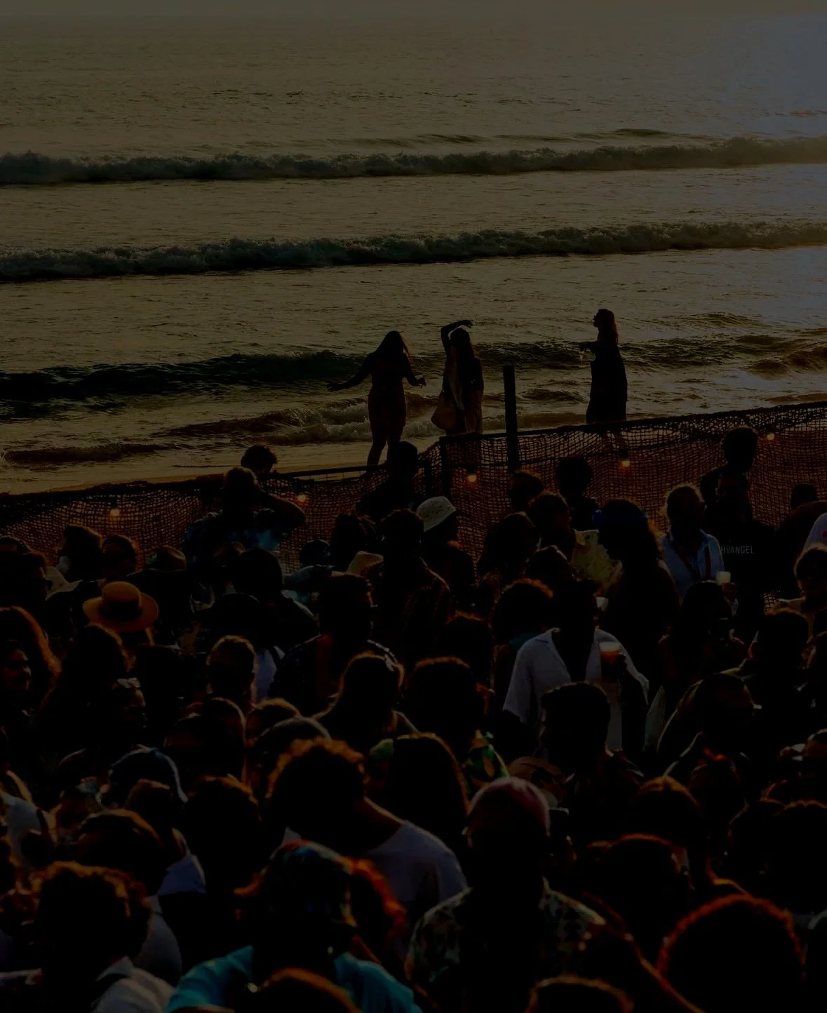 Silhouettes of people on a beach at sunset, with a large crowd in the foreground, some sitting and some standing near the shoreline.