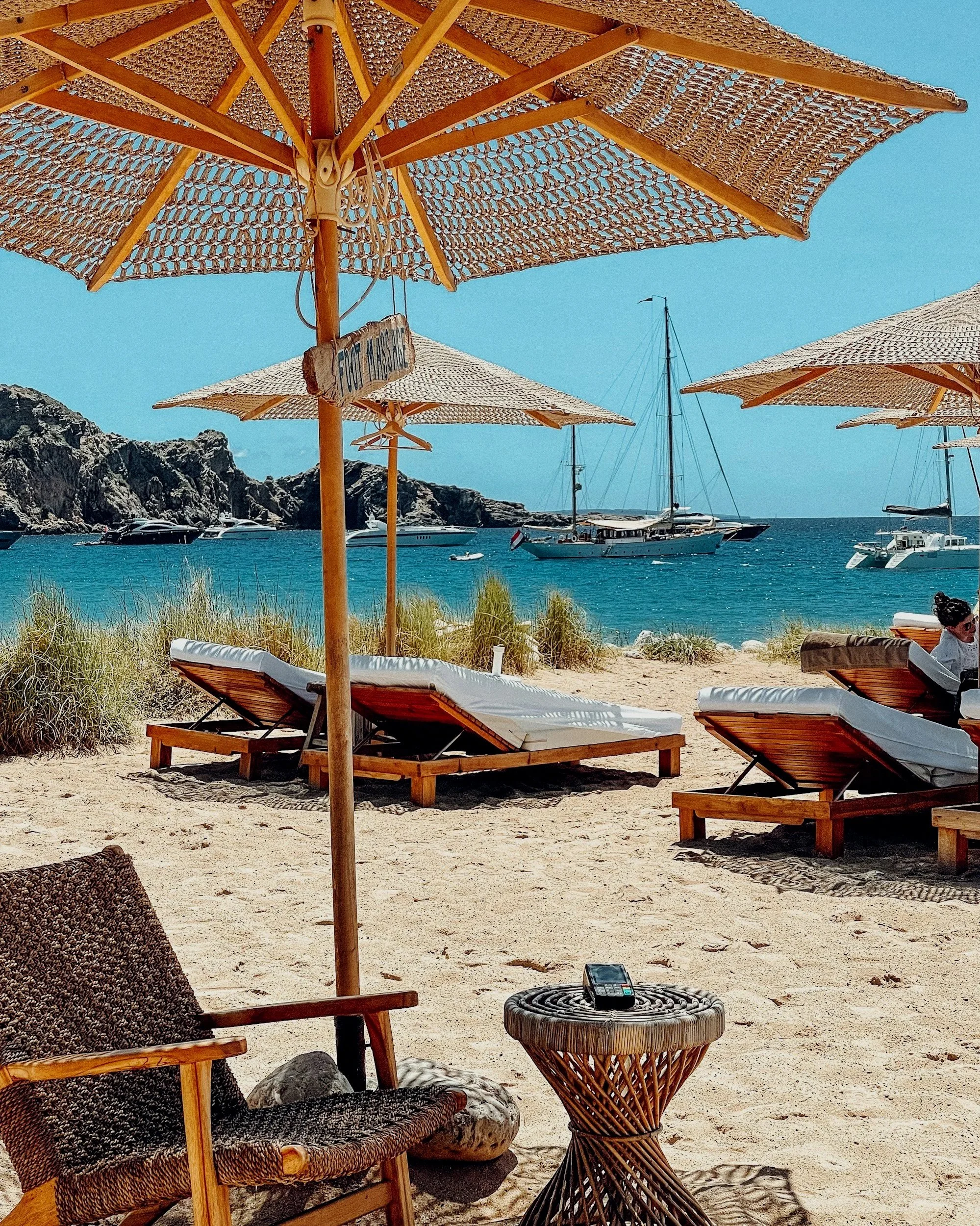 Beach scene with wooden lounge chairs and large woven umbrellas. Several boats are anchored in the water near rocky cliffs, under a clear blue sky.