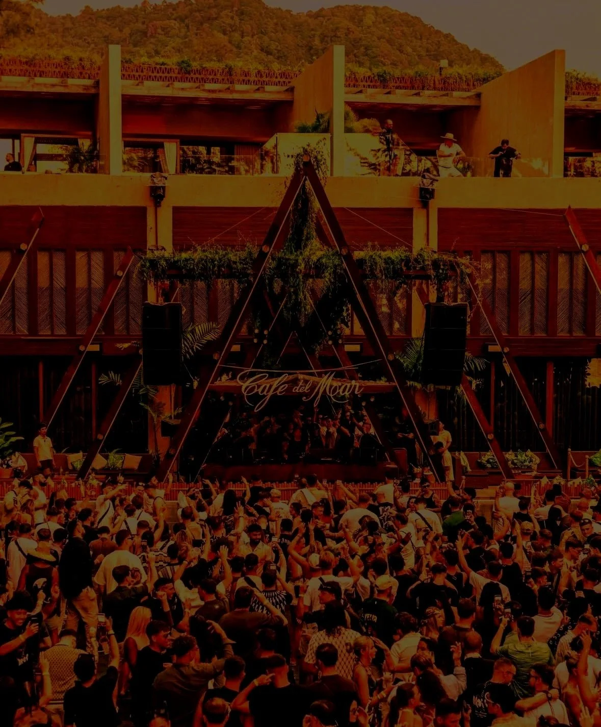 A crowded outdoor event with a stage labeled 'Café del Mar' and people gathered in front, surrounded by a building with balconies and greenery, mountains in the background.