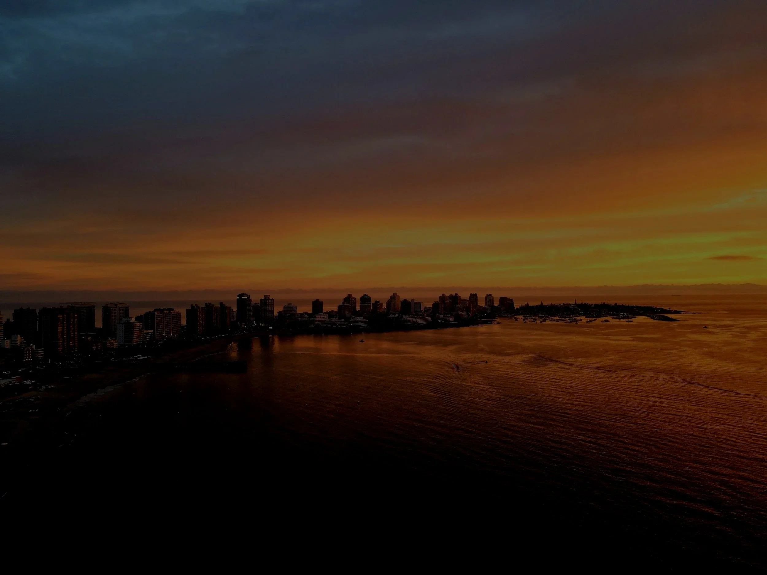 Sunset over a city skyline with high-rise buildings, water in the foreground, and a colorful sky with shades of blue, orange, and yellow.