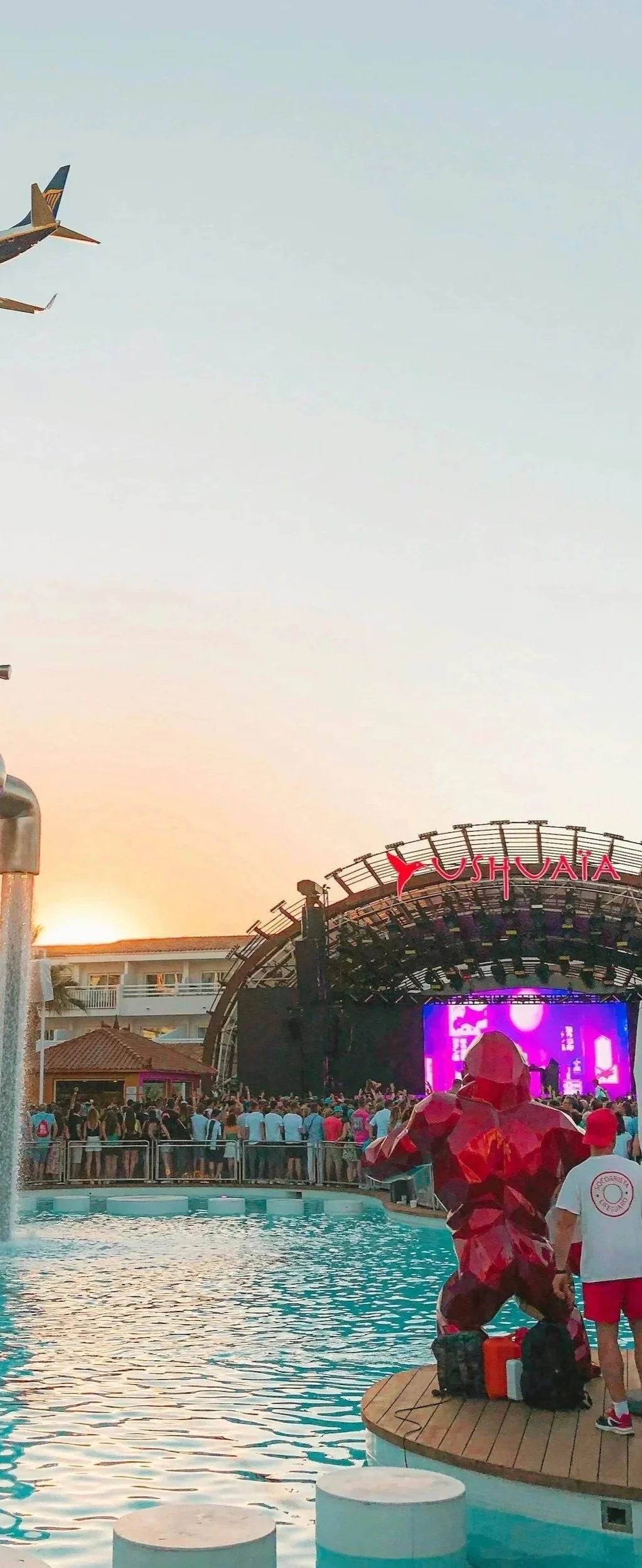 A crowd gathered near a stage at Ushuaia, an outdoor nightclub. There is a swimming pool in the foreground with a red sculpture of a bear nearby. Two airplanes are flying in the sky above the scene during sunset.