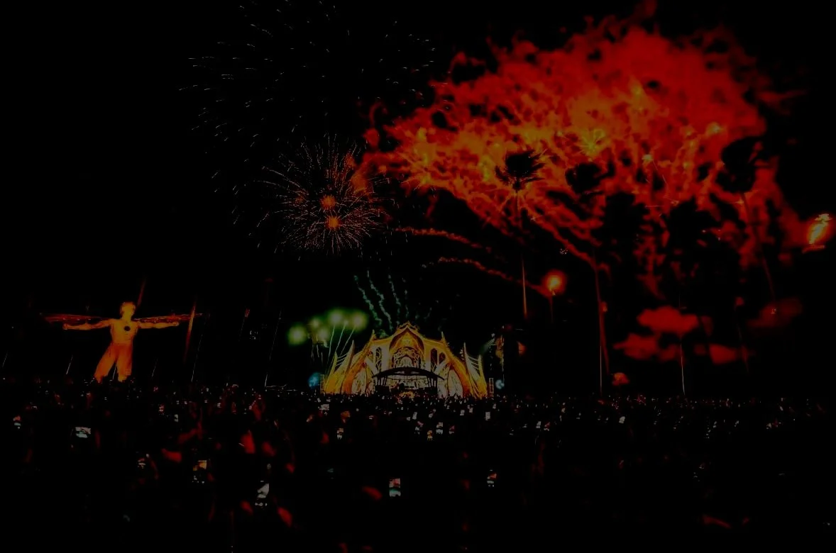 Nighttime scene with fireworks above a concert stage, a statue with arms outstretched on the left, and Aztec-style architecture in the background, with a crowd of people in the foreground.