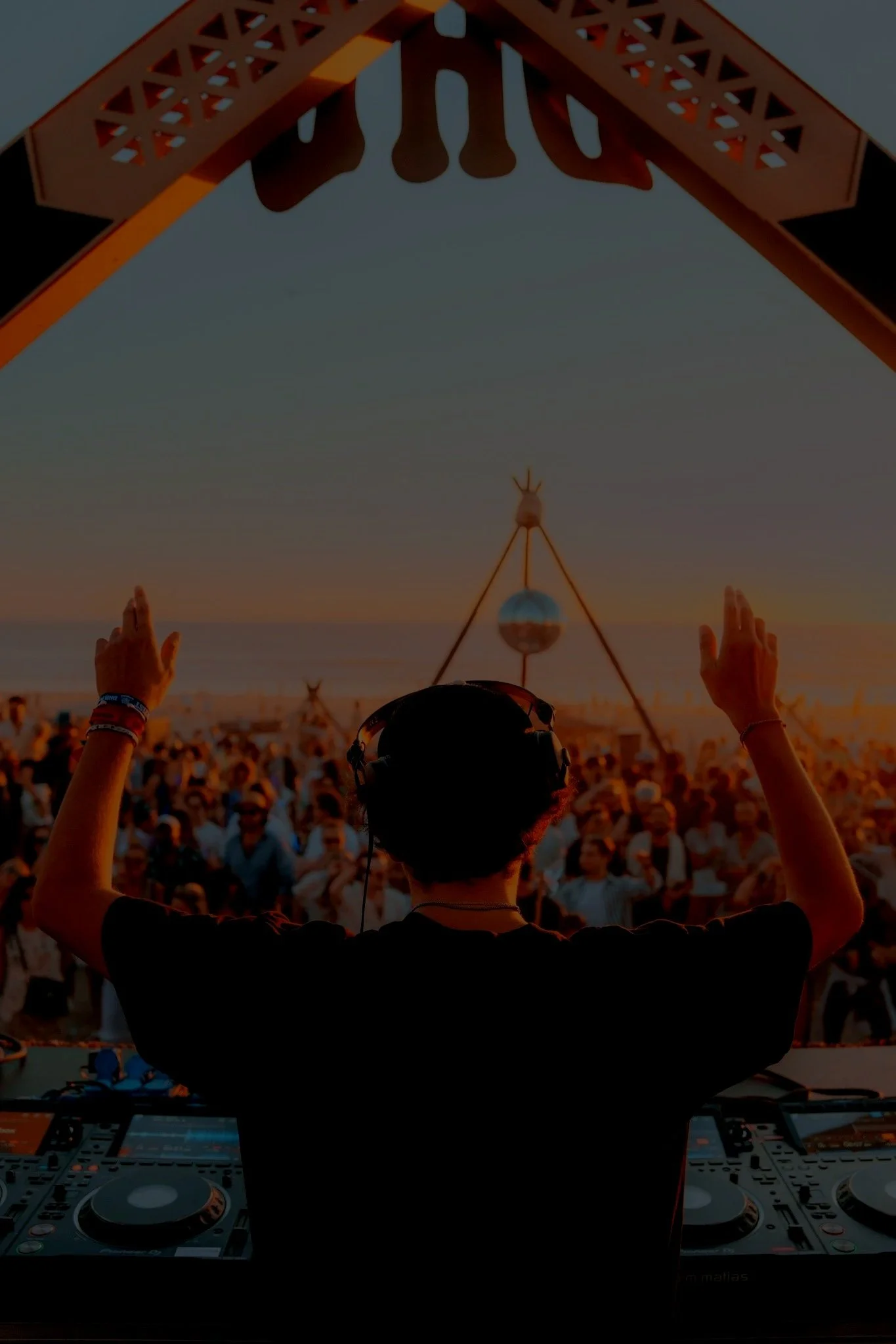A DJ performing at an outdoor music festival during sunset, with a large crowd watching.