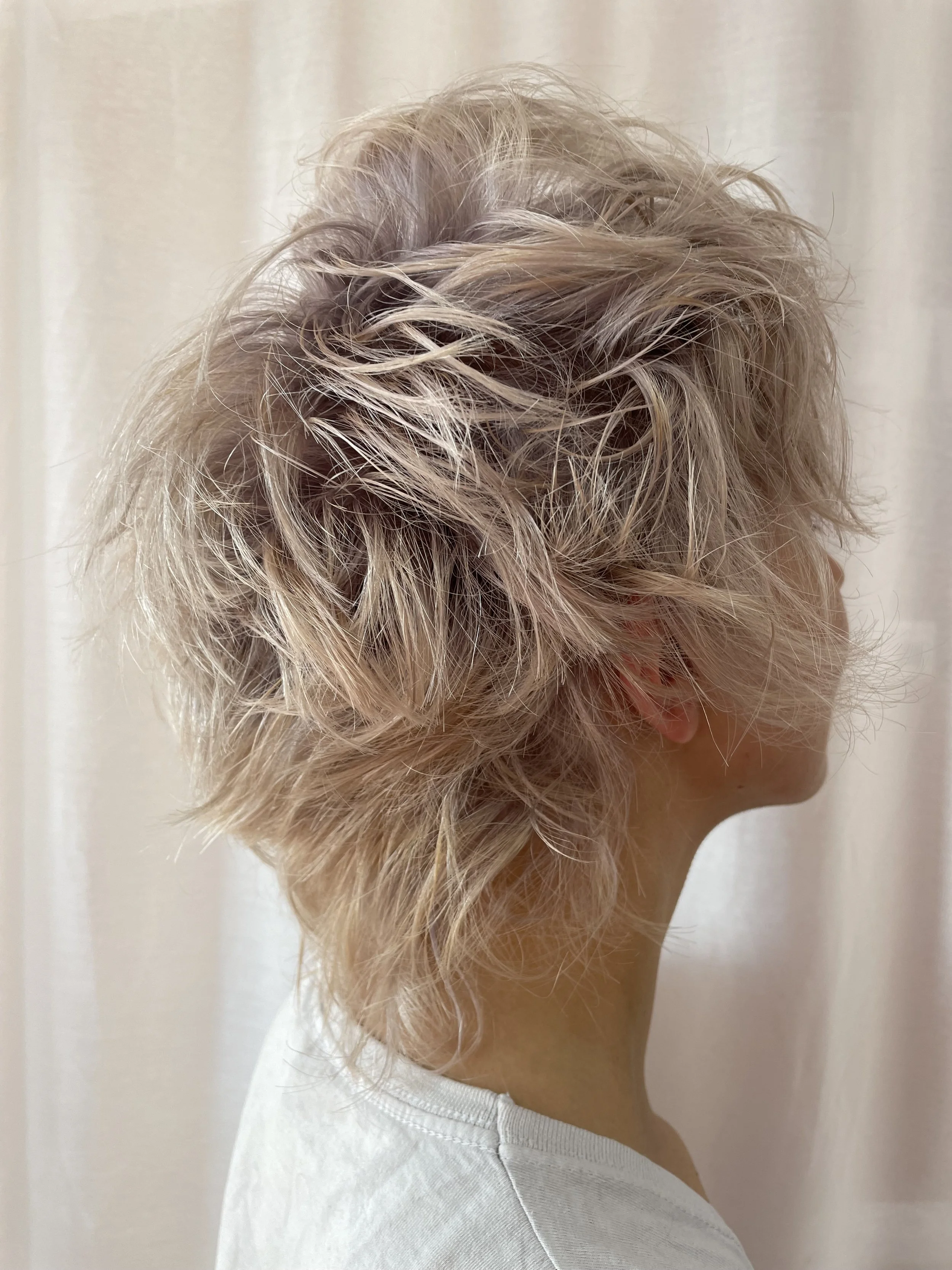 Side view of a person with short, curly, platinum blonde hair wearing a white top.